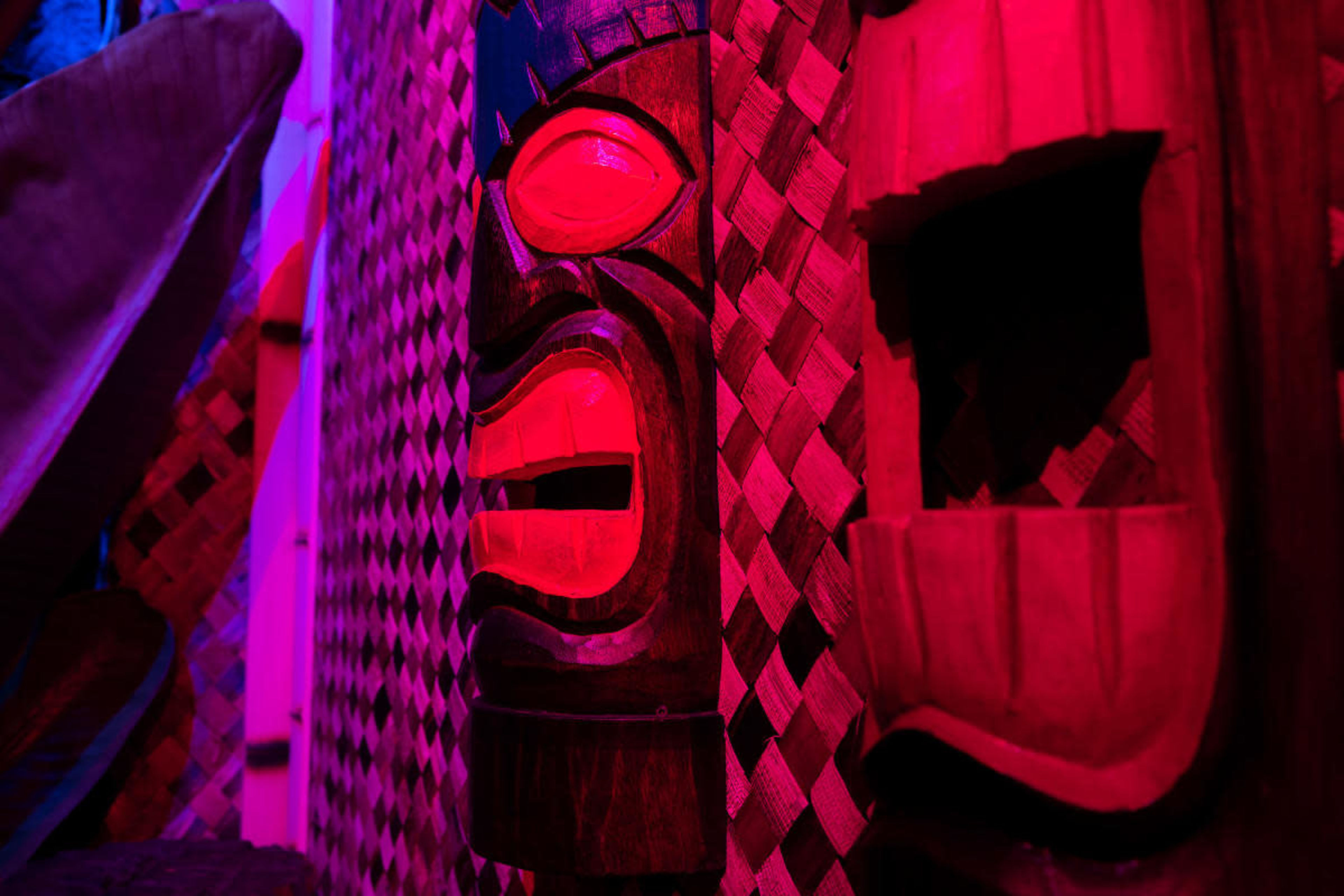 The image features a carved wooden mask with red accents, displayed against a textured background of woven material.