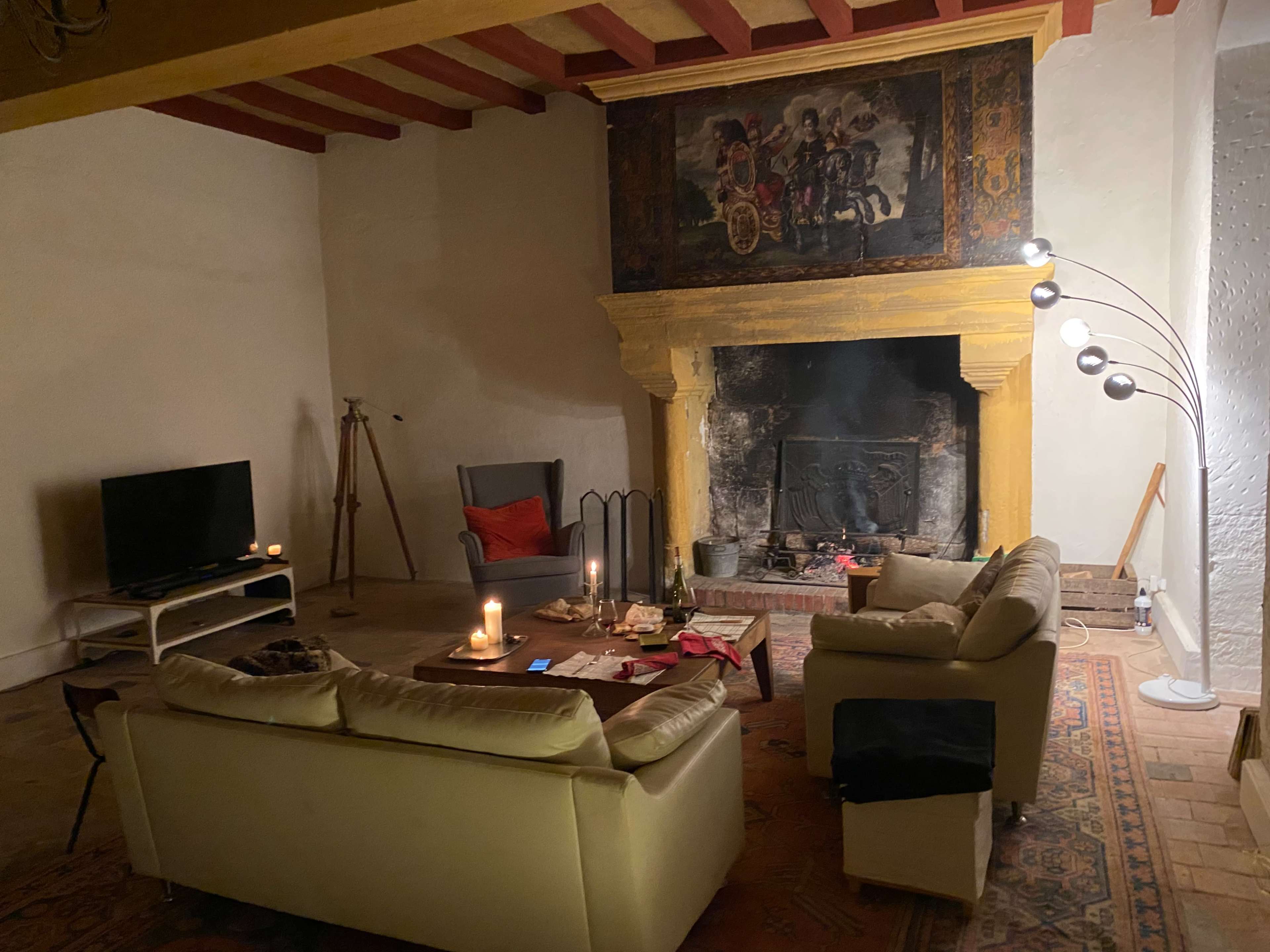 The image shows a living room featuring a yellow-painted fireplace, two sofas, a television, and a floor lamp, with candles arranged on a coffee table.