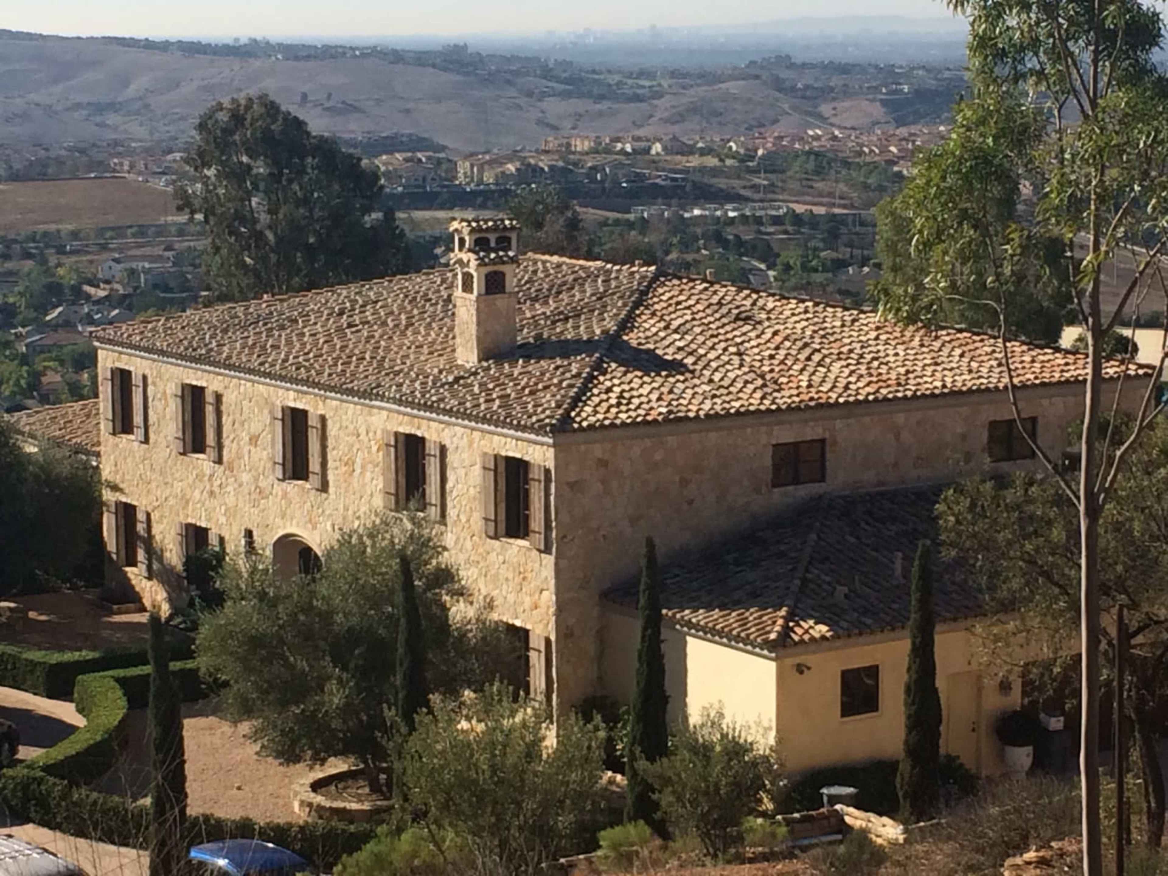 A large stone house with a tiled roof is situated on a hillside, surrounded by greenery and valleys.