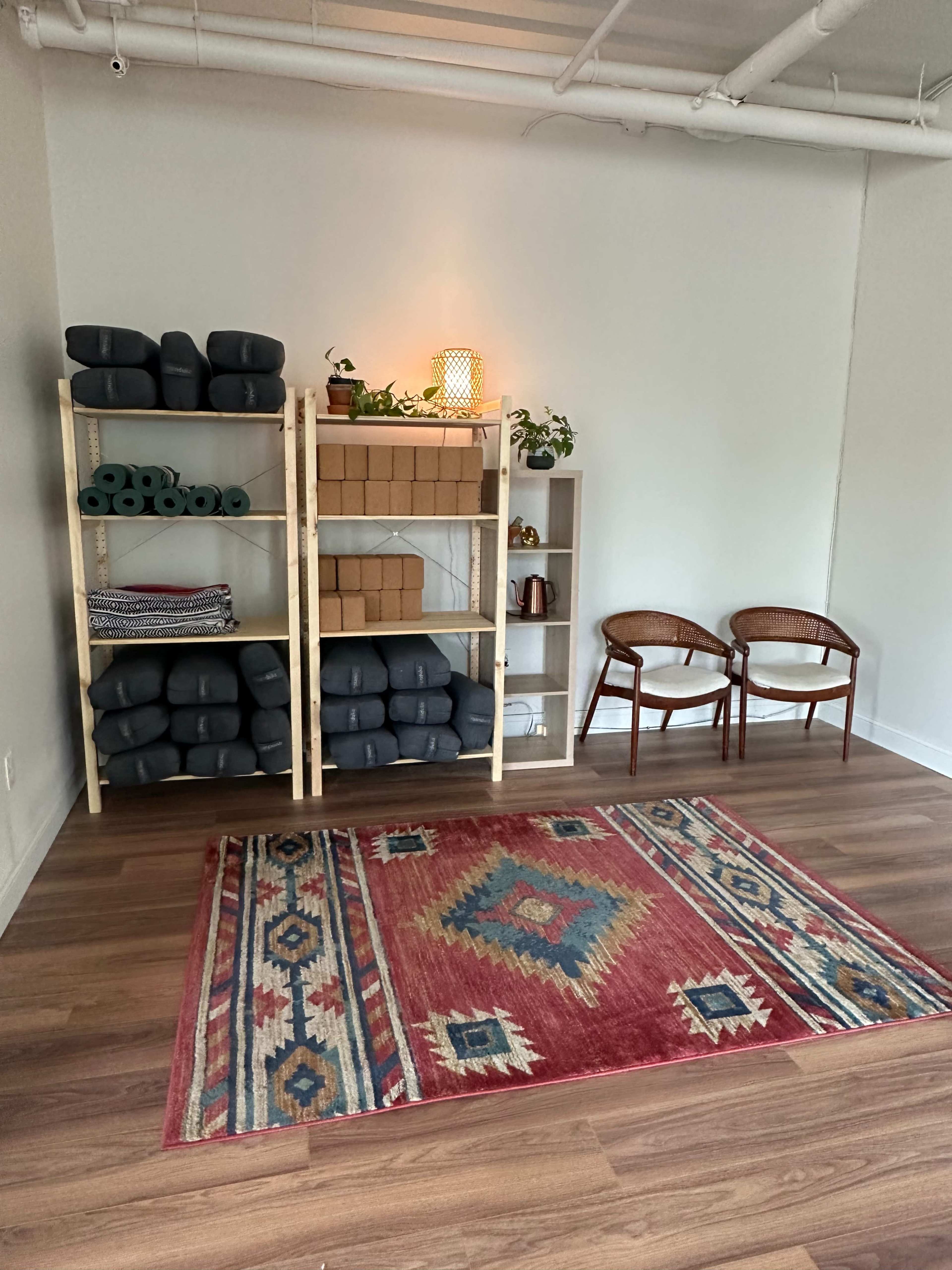 The image shows a minimalist interior space with two wooden chairs, a small shelf containing yoga mats and blocks, and a colorful rug on the floor.