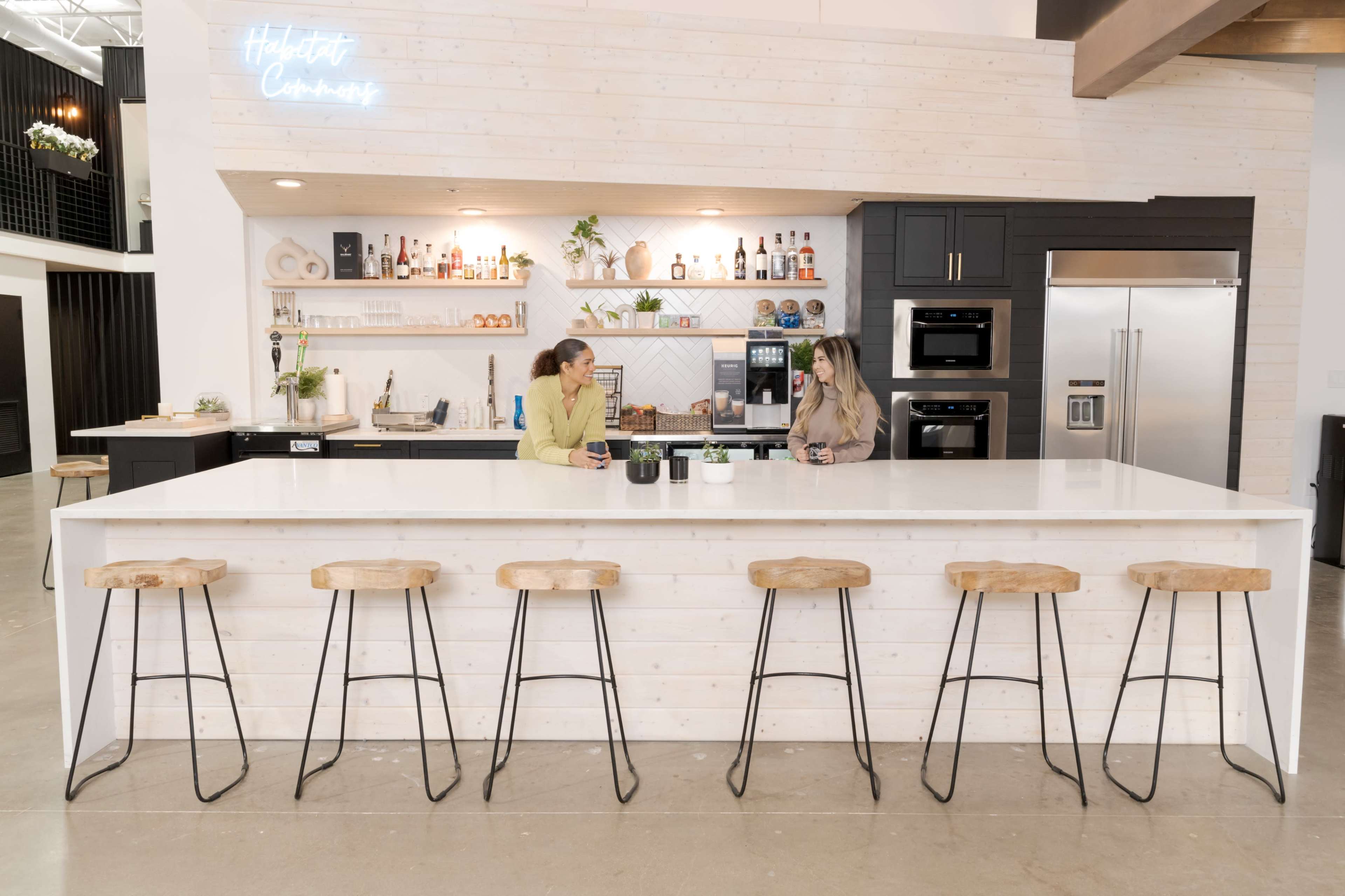 Two women are seated at a kitchen island with wooden stools, engaged in conversation, while a modern kitchen with a light-colored wooden wall and dark cabinetry is visible in the background.