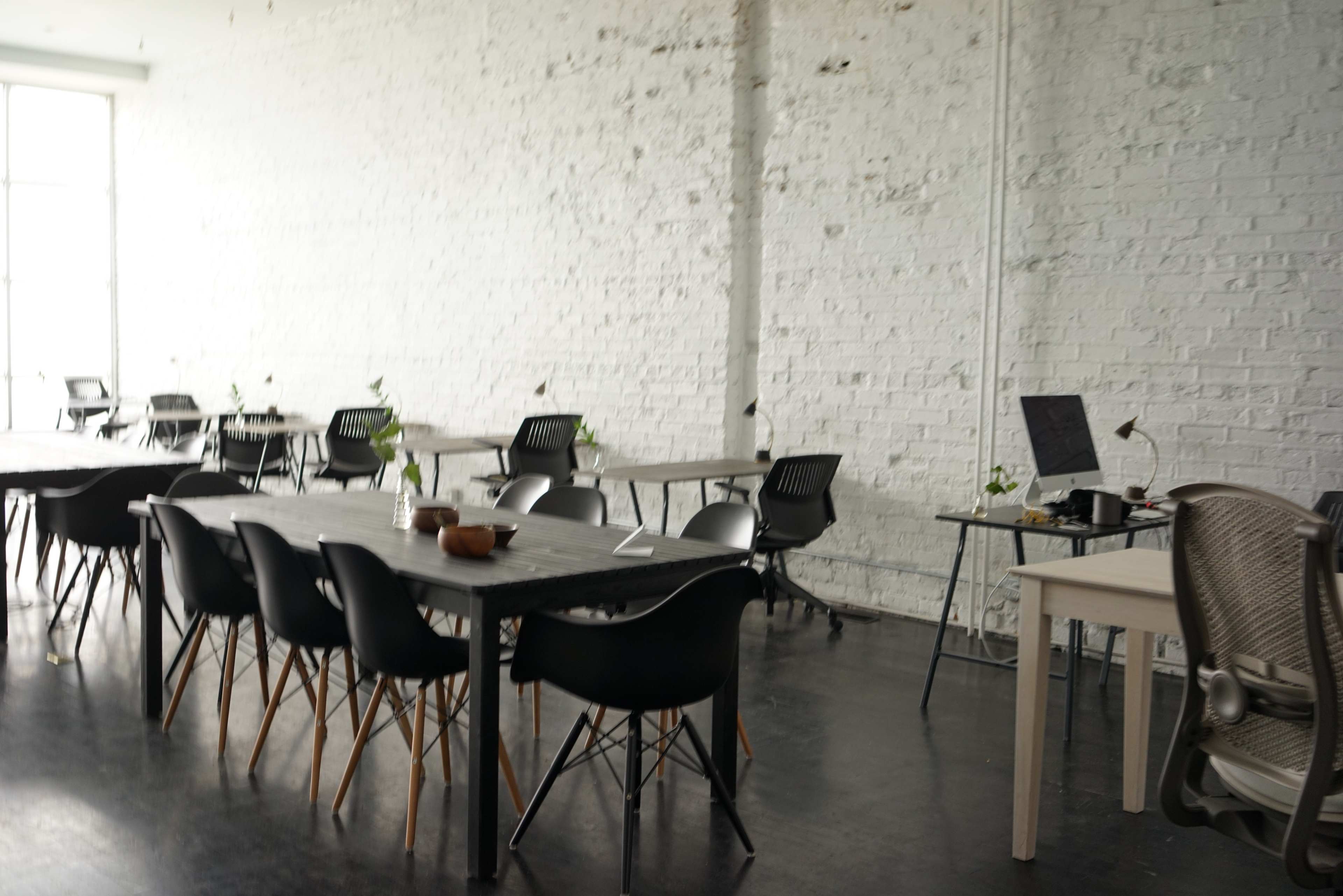 A spacious office interior features several black and white tables with chairs, a computer setup, and small plants against a white brick wall.