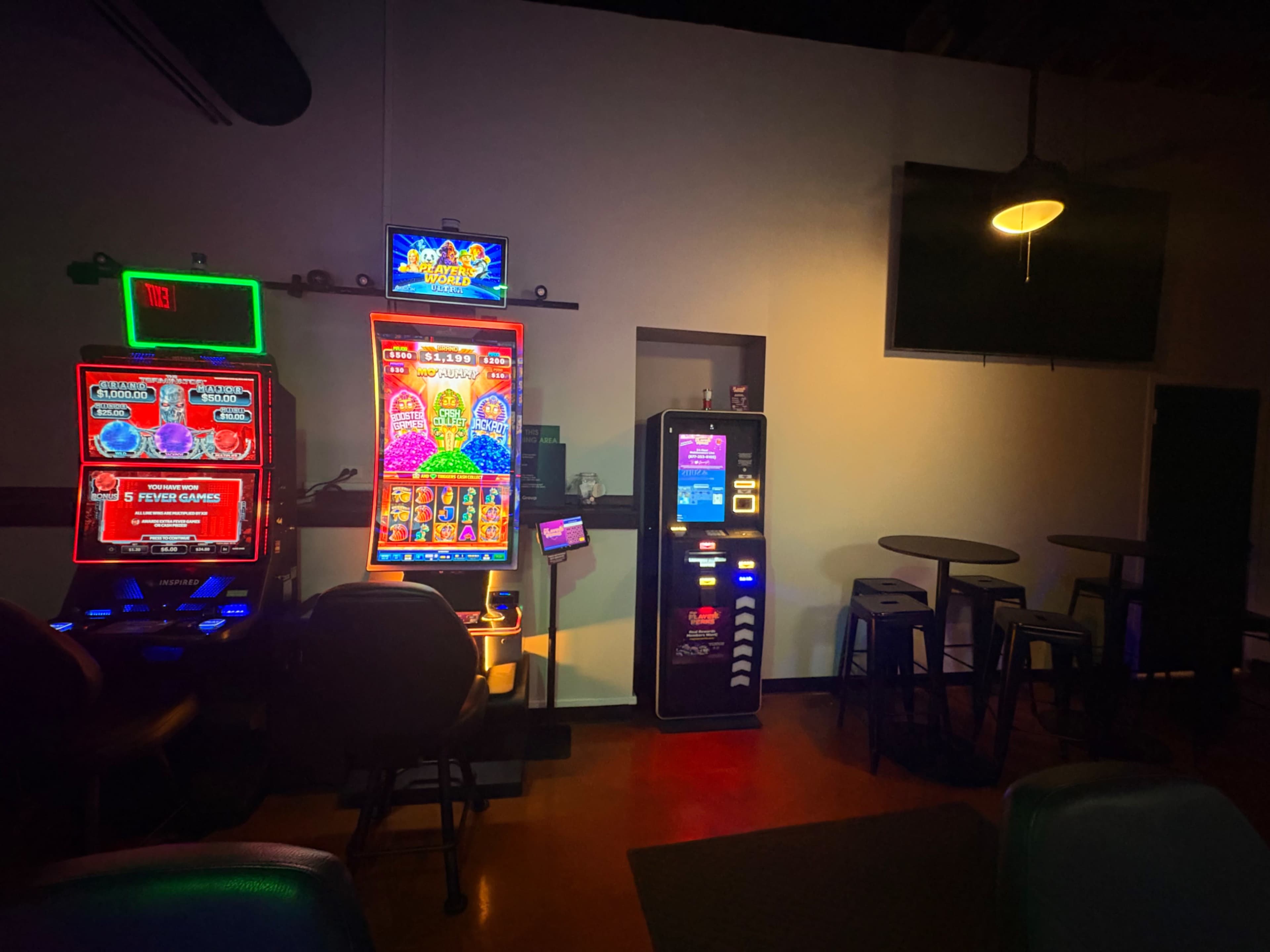 The image shows a dimly lit gaming area featuring three slot machines and a few empty tables.