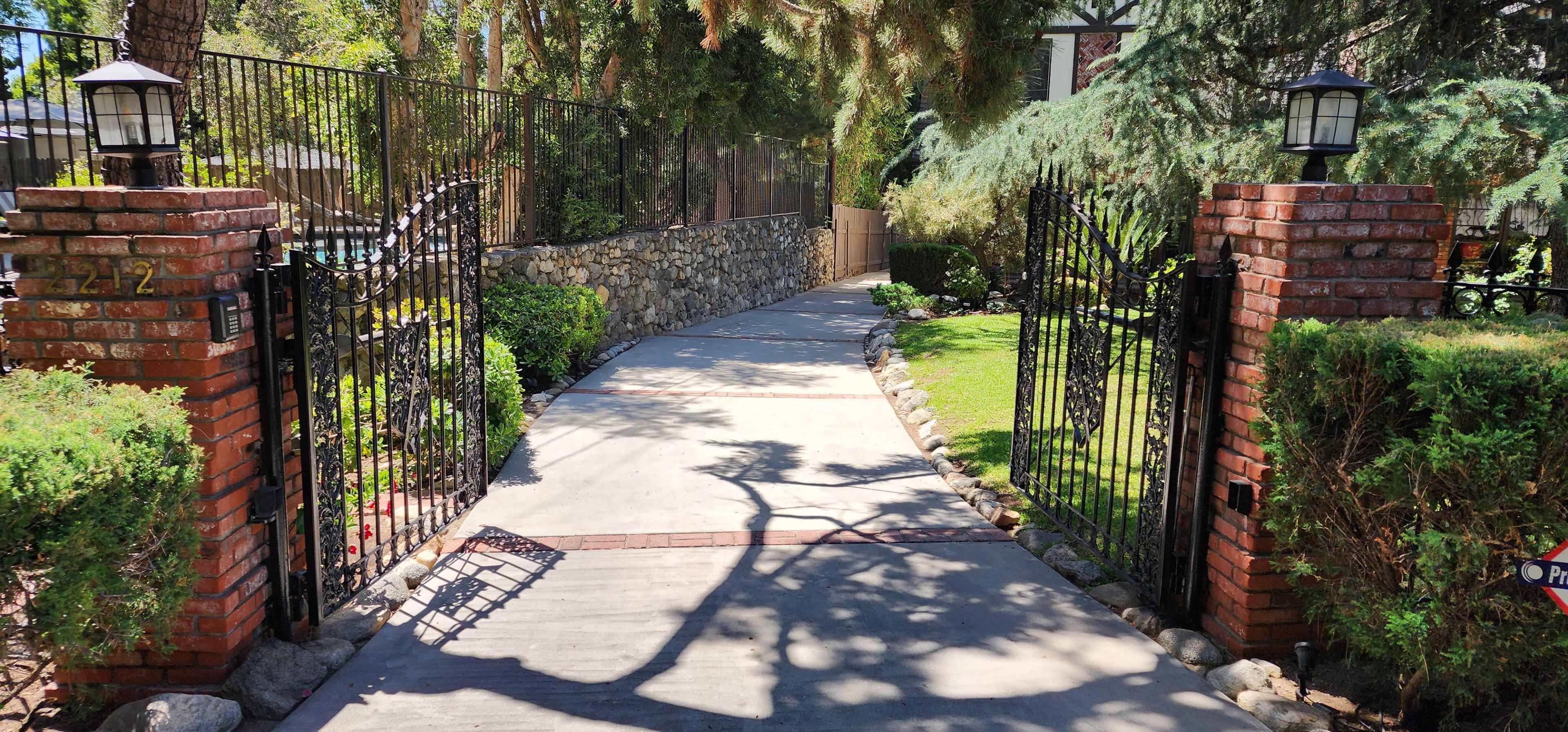 A paved walkway leads through ornate black gates, flanked by brick pillars and lush greenery, toward a house in the background.