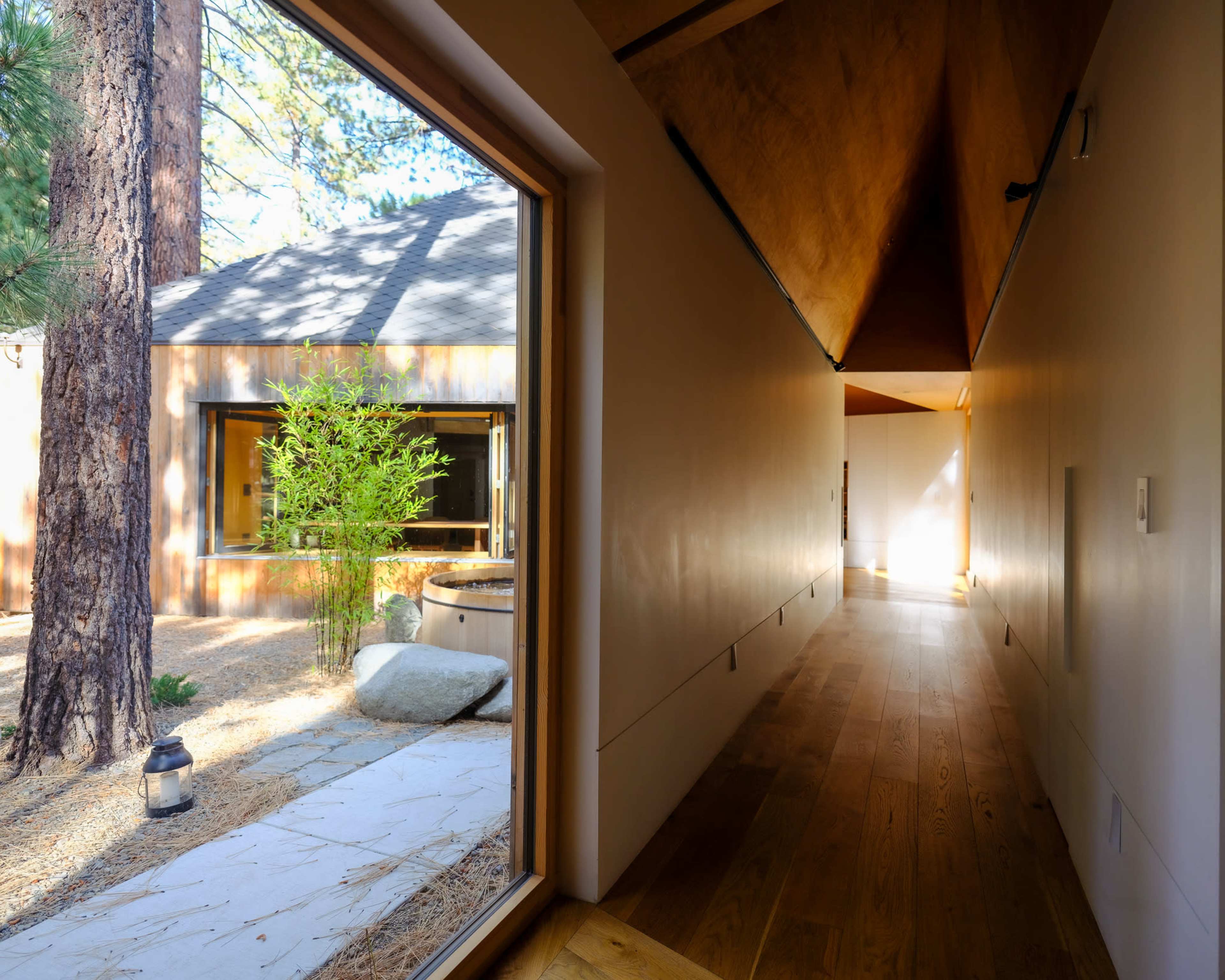 A narrow hallway with wooden flooring leads to a bright open space, framed by a large window showing a tranquil outdoor area with trees and rocks.