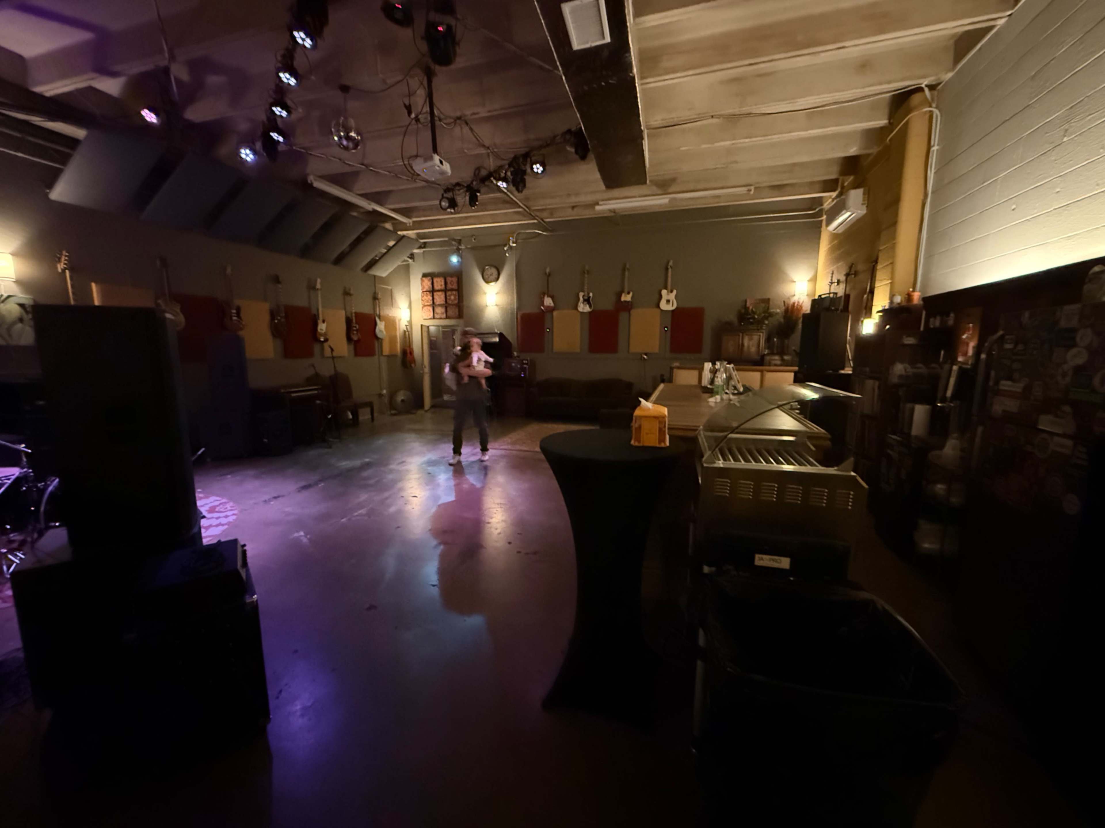 The image shows a spacious rehearsal studio with multiple guitars hanging on the walls and a performer standing in the center under stage lights.