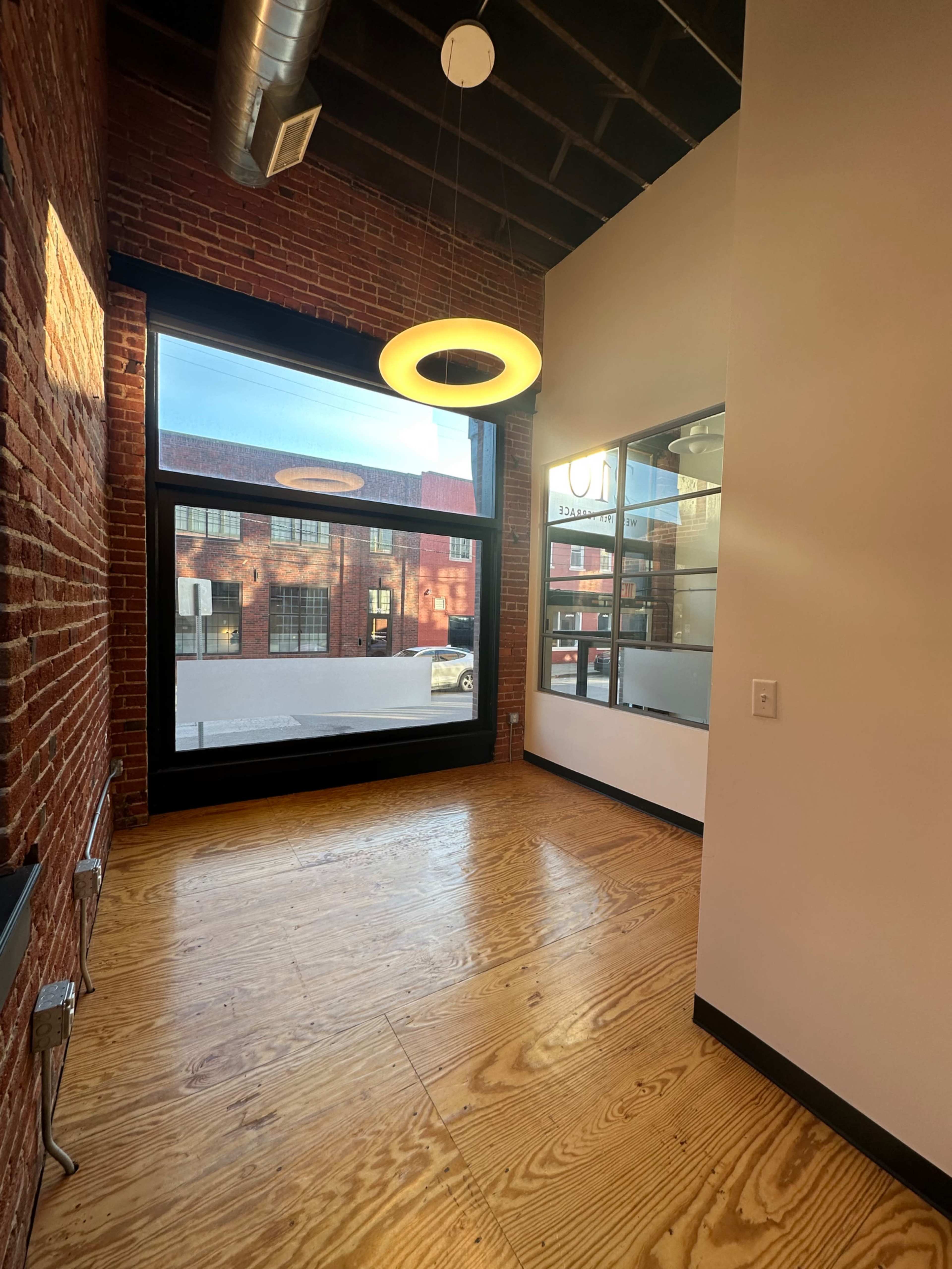 The image shows an interior space featuring exposed brick walls, wooden floors, and a large window with a circular light fixture hanging from the ceiling.