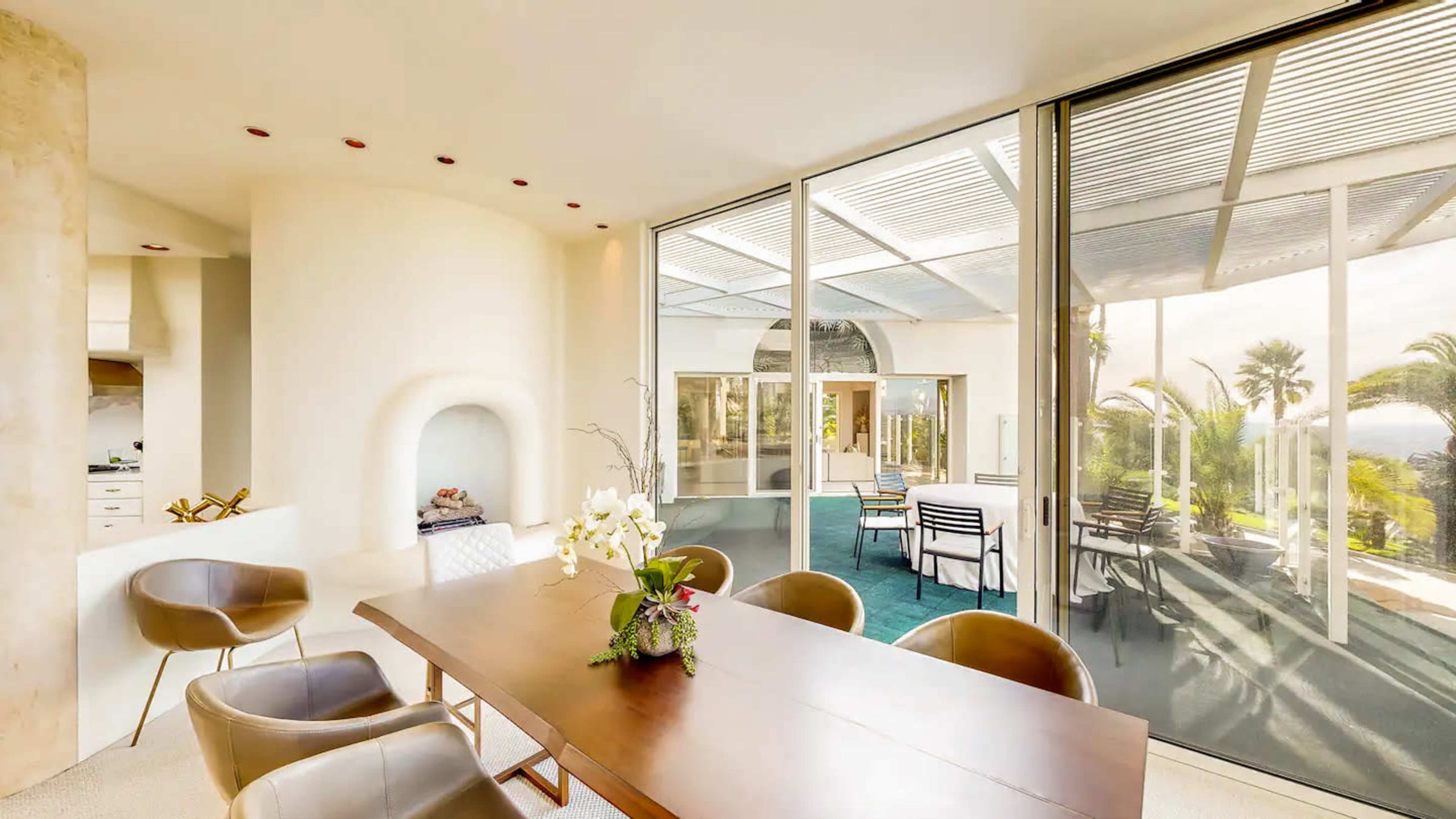 A modern dining area features a large table surrounded by chairs, with large glass windows opening to a patio view of palm trees.