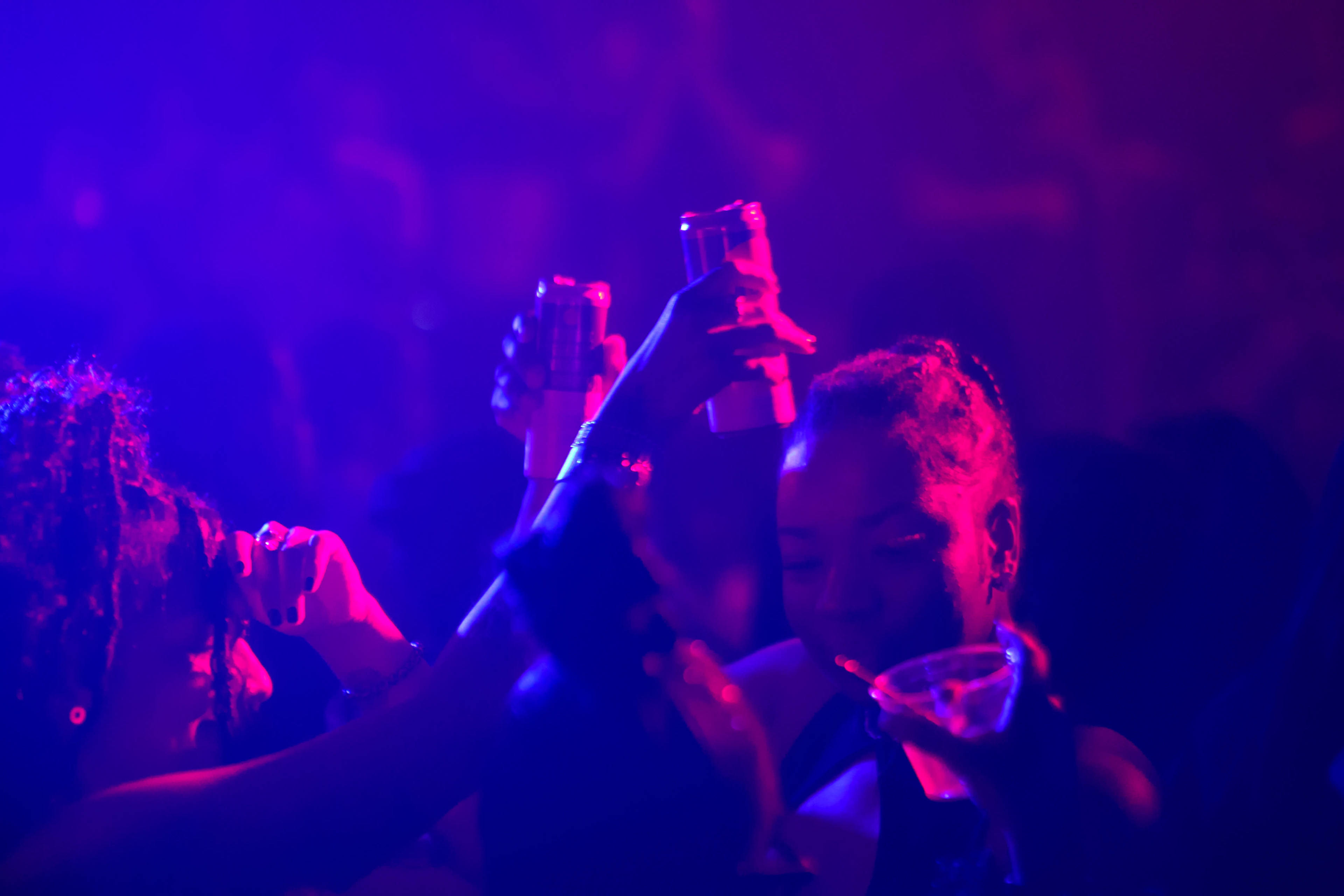 A group of people are raising their drinks in celebration at a dimly lit party with colorful lighting.