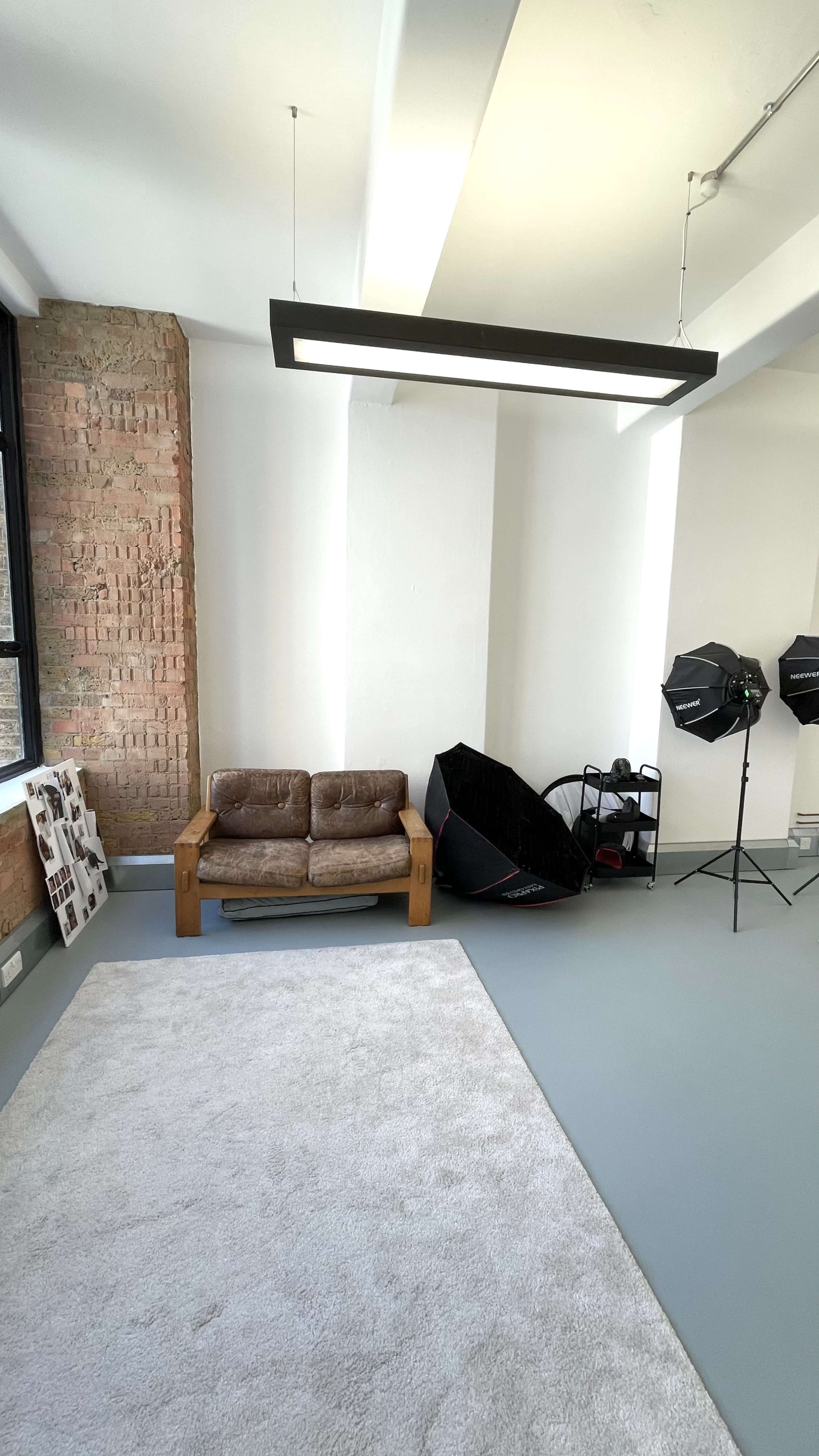 The image shows a spacious room with a wooden sofa, a large light fixture, a white rug, and photography equipment in the corner.