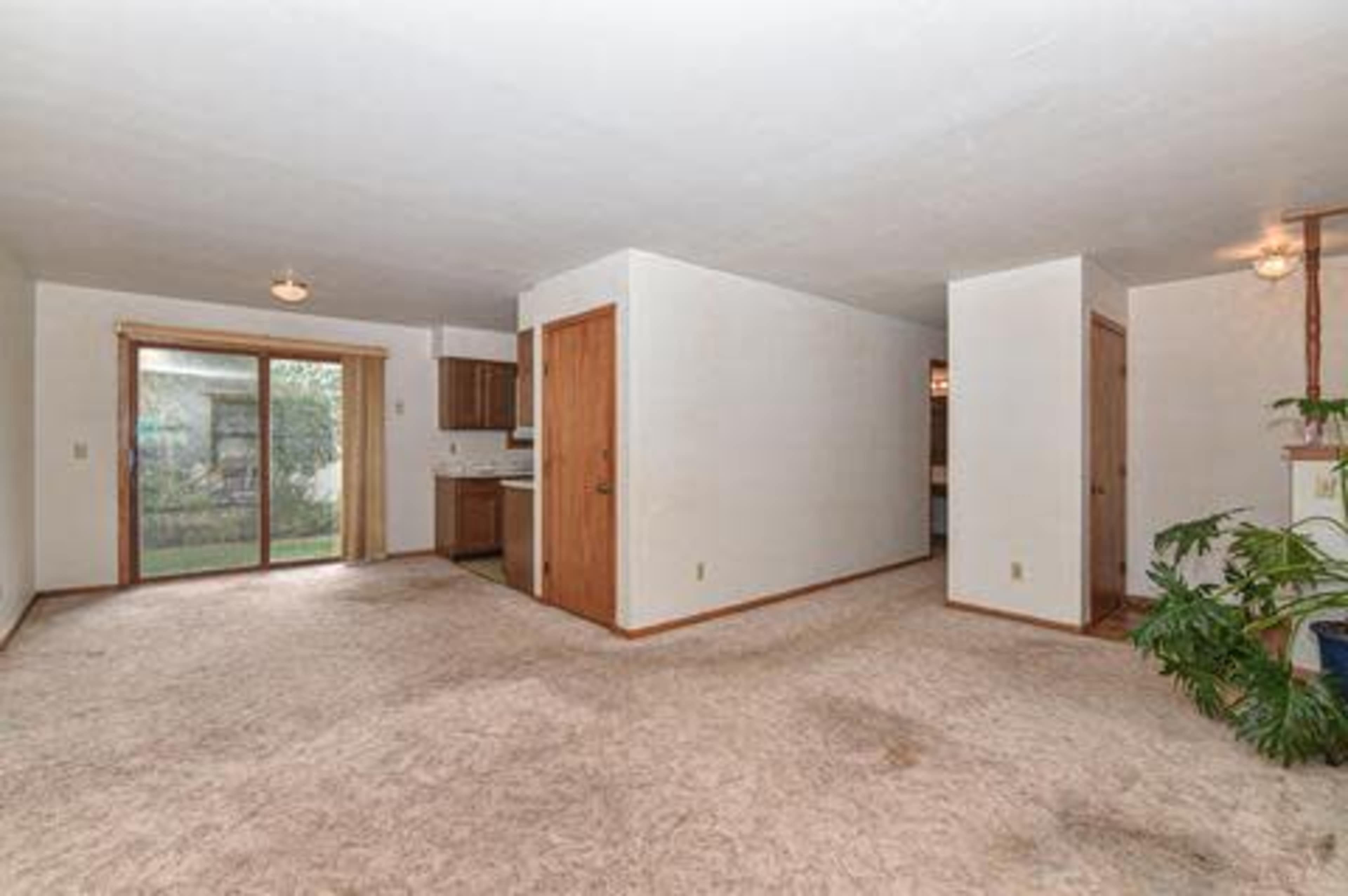 An empty living space with carpeted flooring, including a kitchen area and a sliding door leading to an outdoor space.