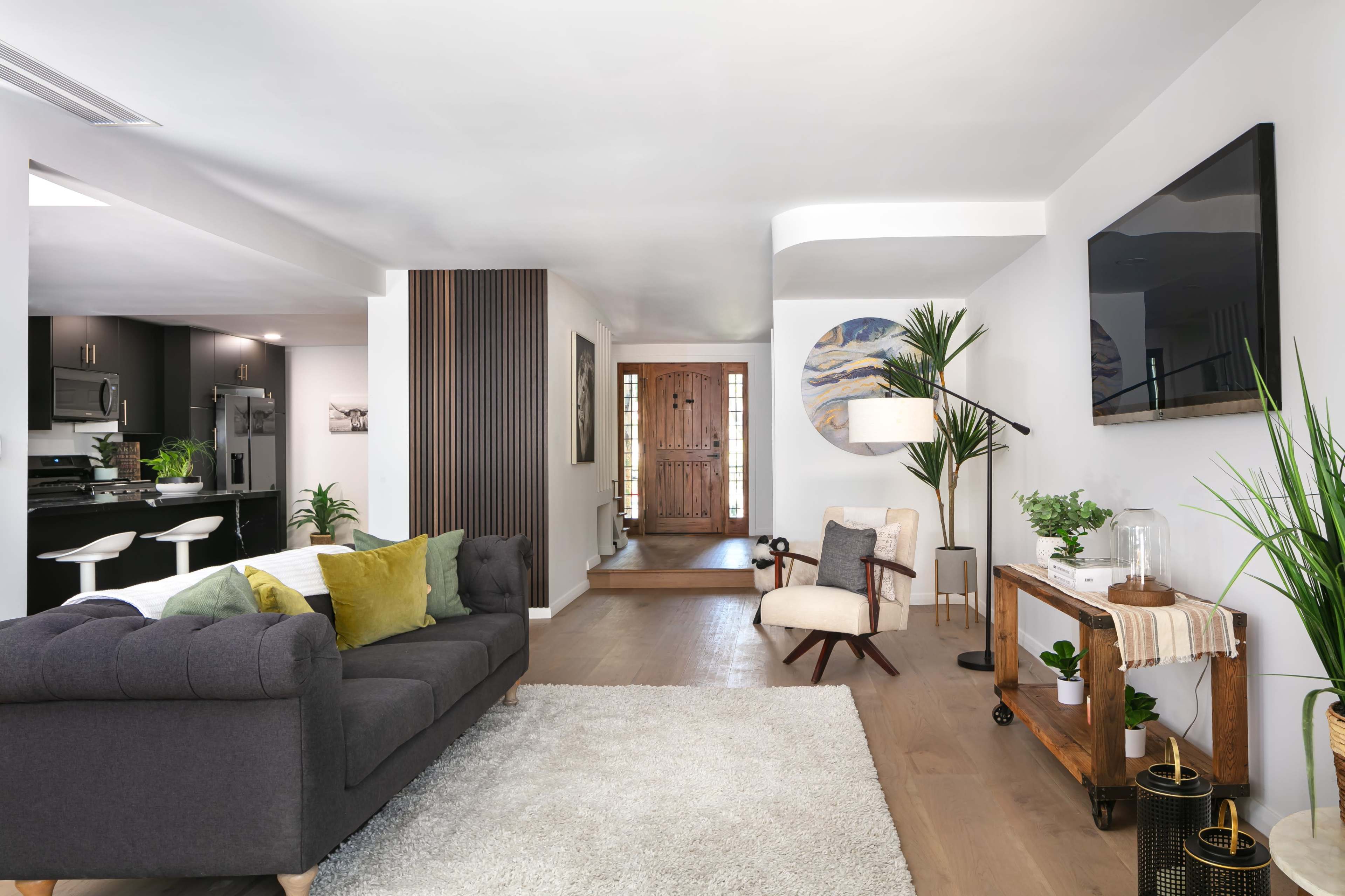 A modern living room features a gray sofa with decorative pillows, a wooden side table, a textured rug, and a view of an entryway with a wooden door and staircase.