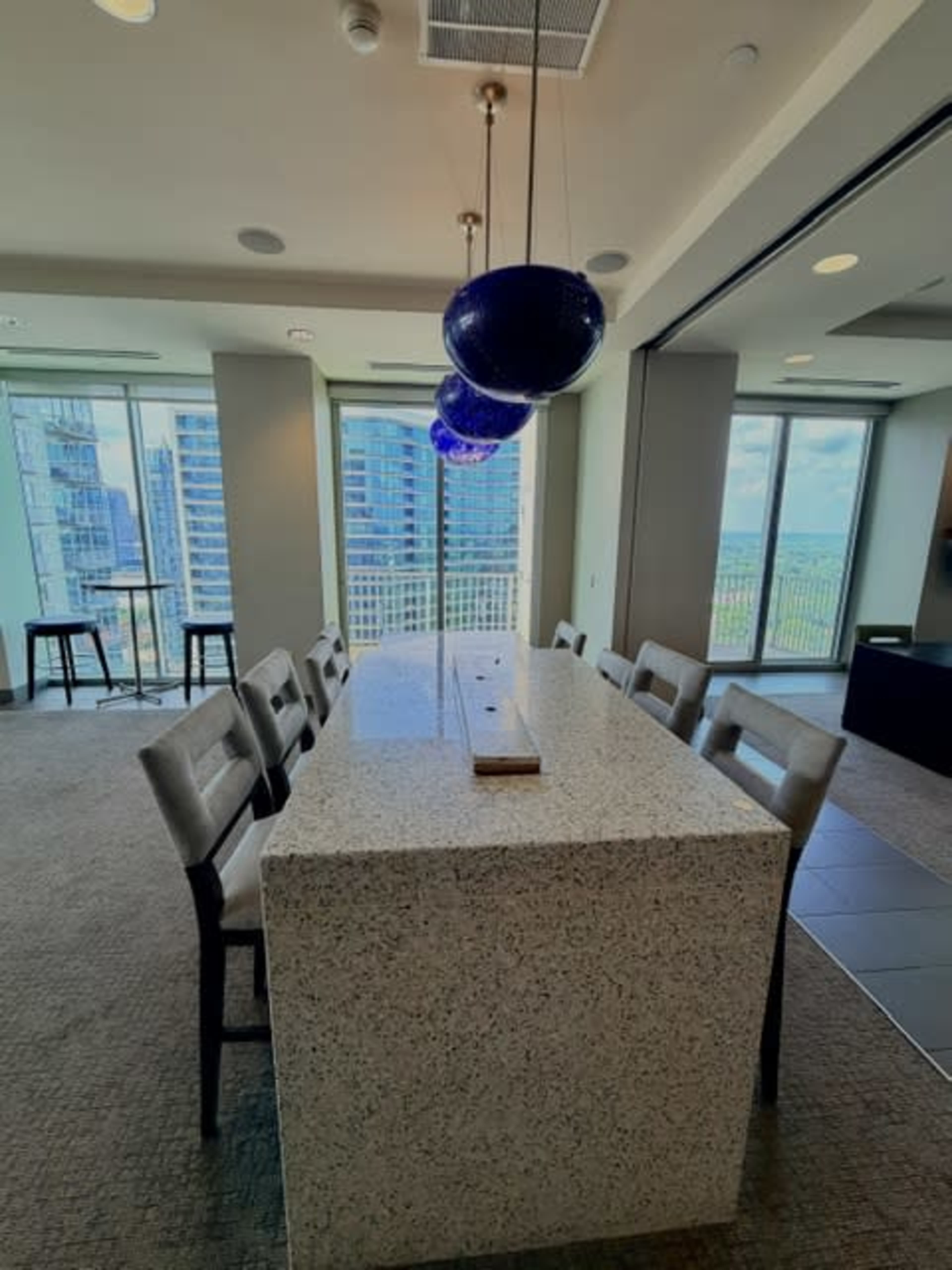 A modern dining area features a large, white speckled table with grey chairs and pendant lights above, all set against a backdrop of large windows showcasing a city view.