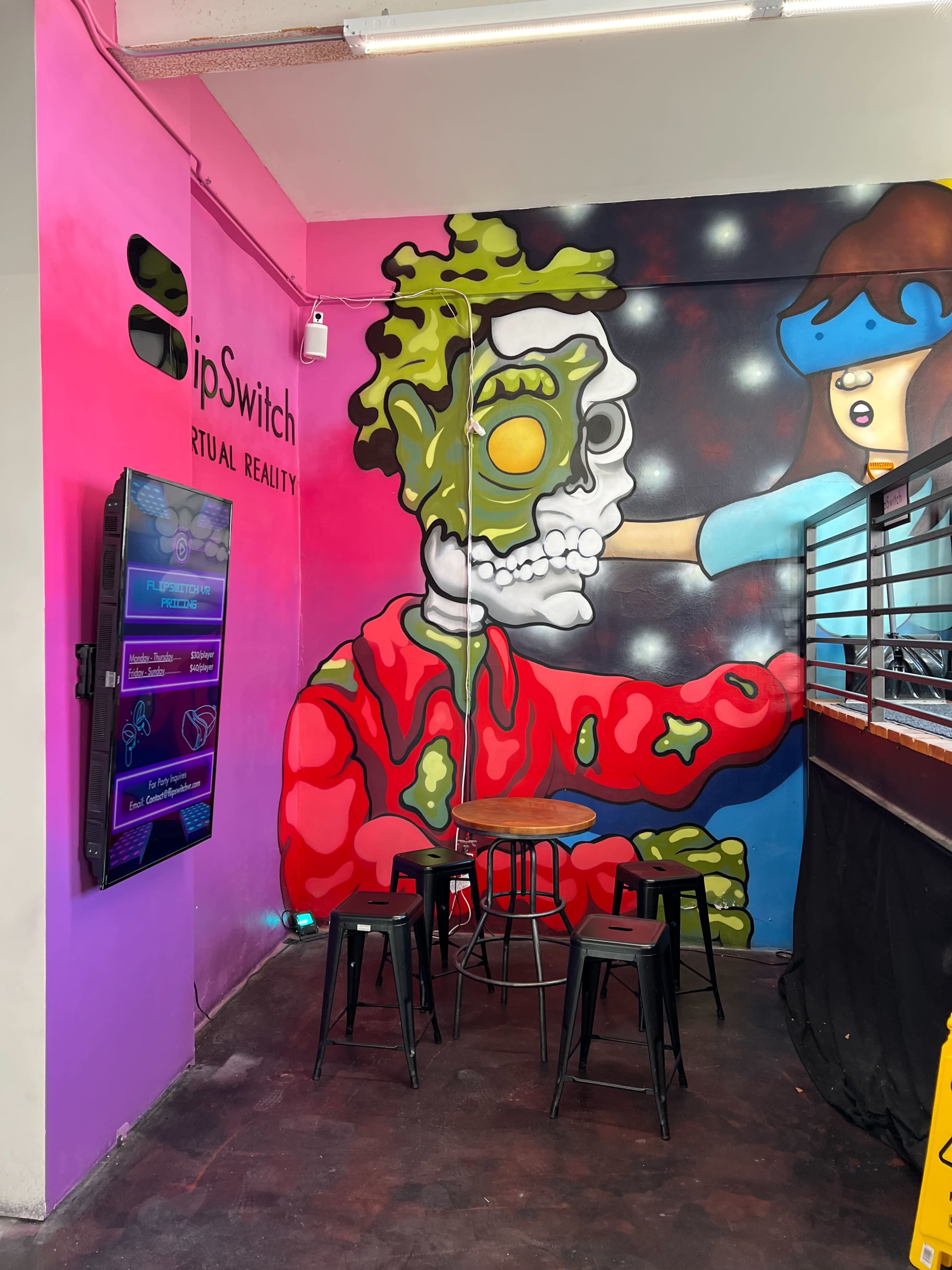 A colorful mural depicting a zombie character and a girl, with a small seating area featuring four black stools and a round table.