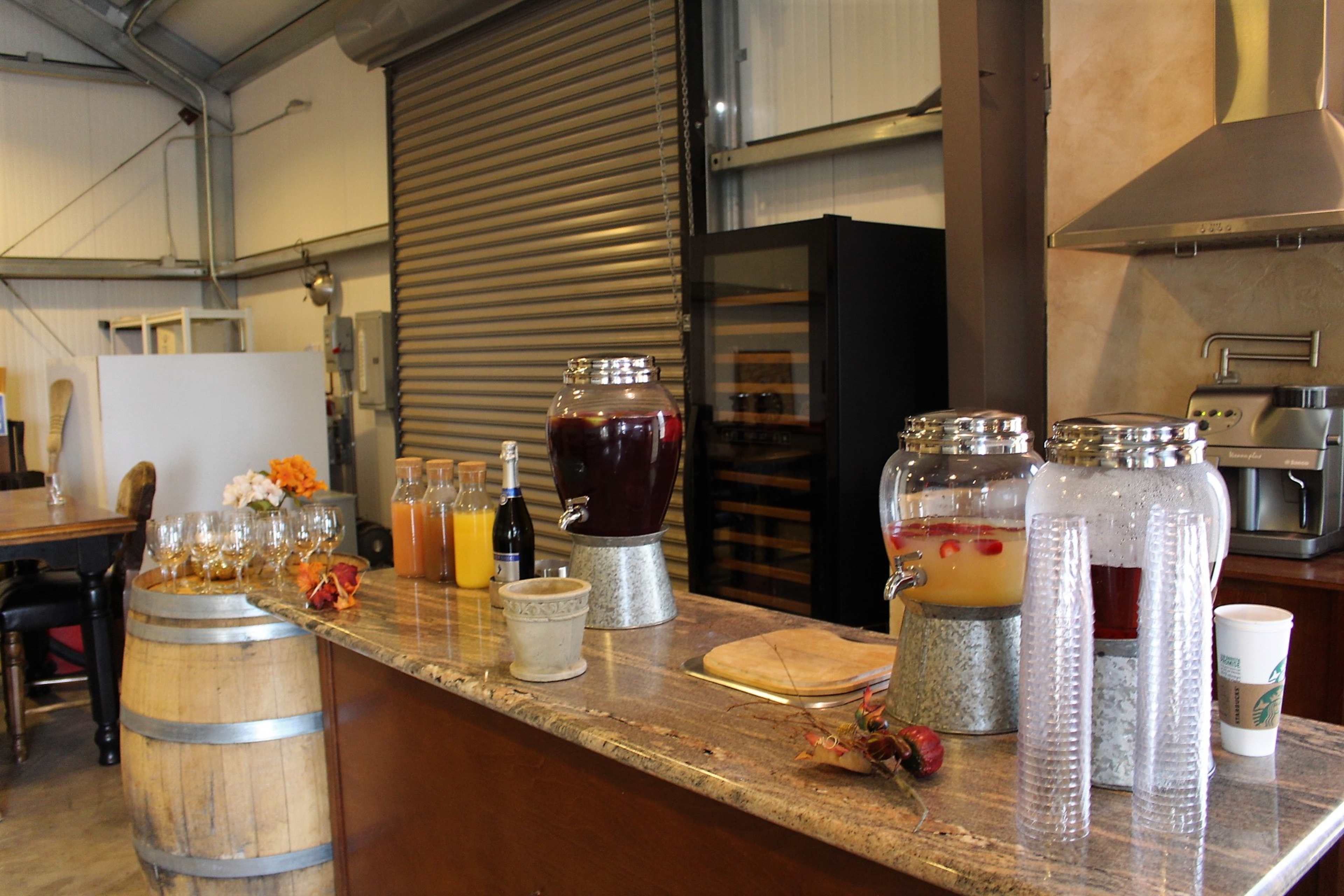 A countertop features a selection of beverage dispensers filled with drinks, along with glasses, fruit garnishes, and a decorative barrel.