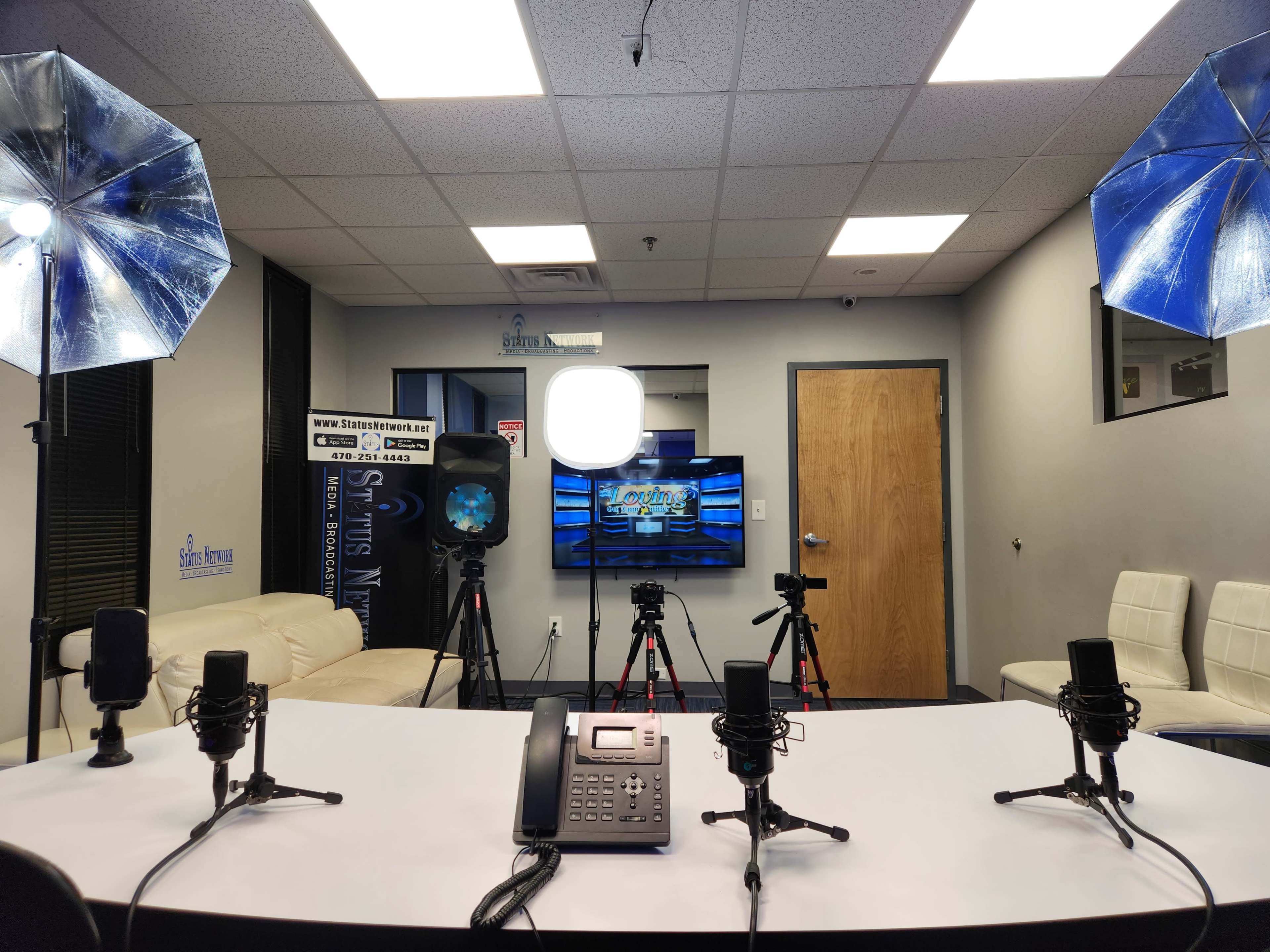 The image shows a video production studio with a table at the center, equipped with microphones and cameras, along with lighting setups and a monitor displaying graphics.