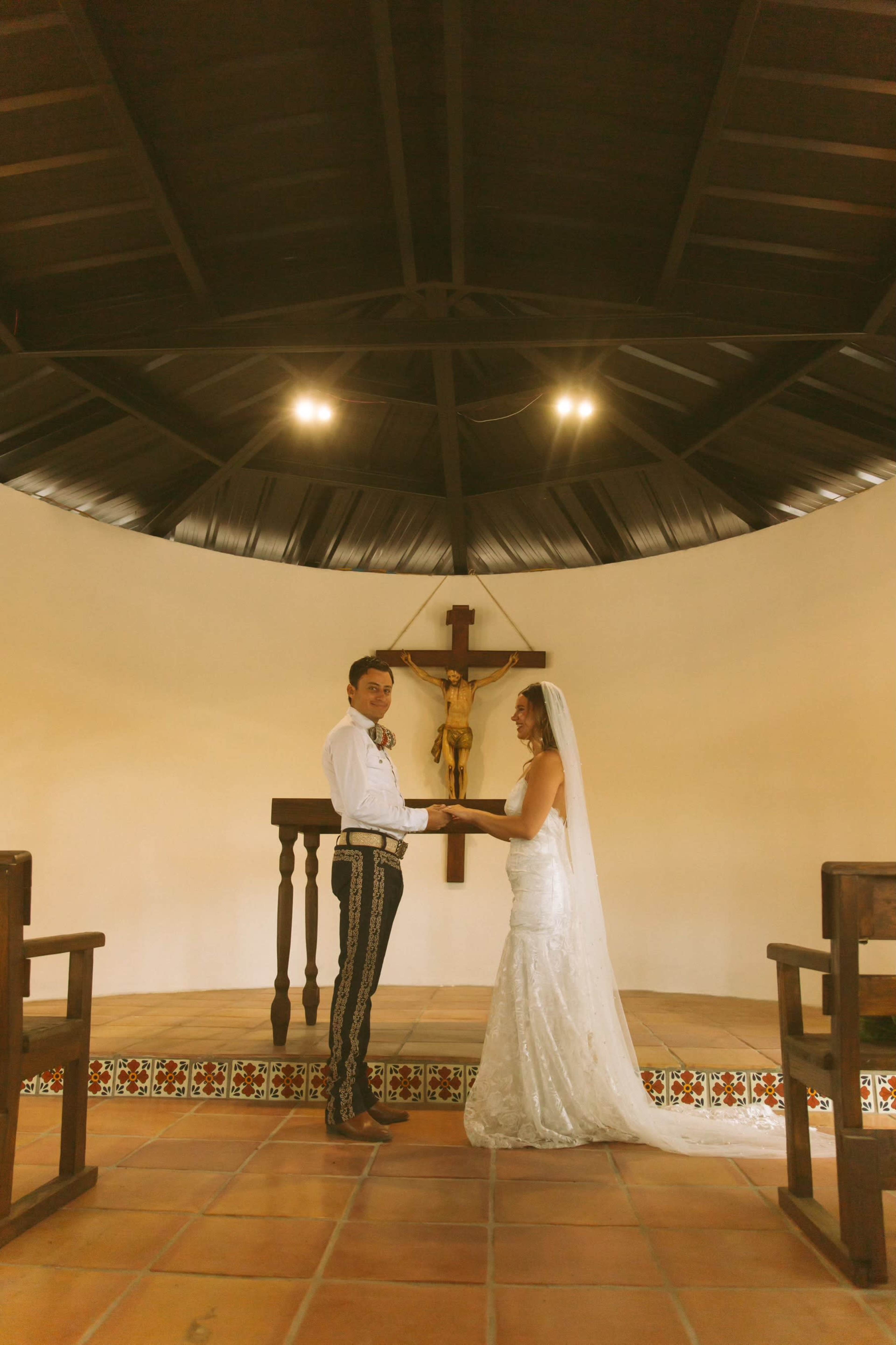 A couple stands facing each other at the altar in a circular chapel, with a crucifix above them and wooden benches on either side.