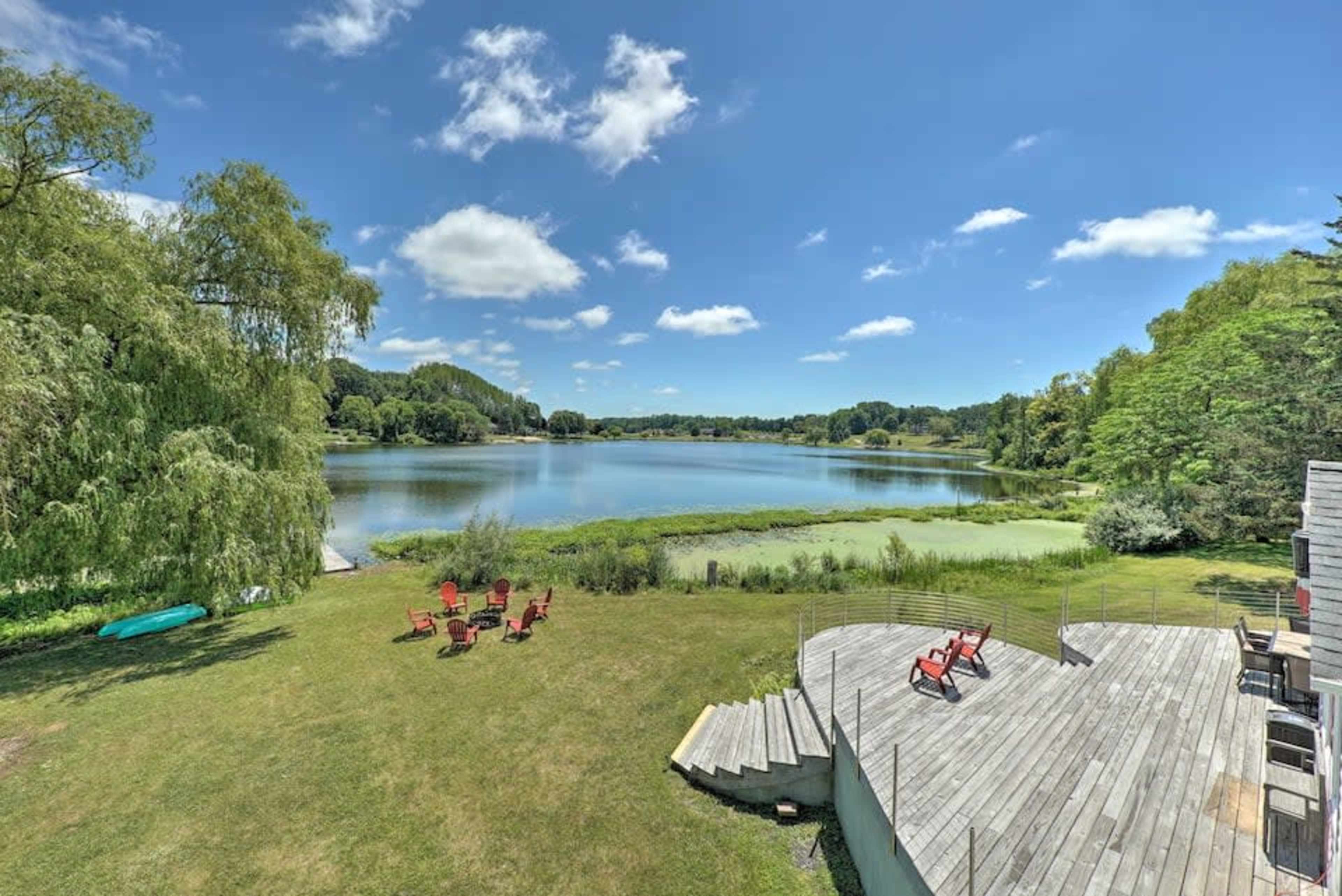 A serene lake surrounded by greenery, with a wooden deck and several red chairs positioned on the lawn.