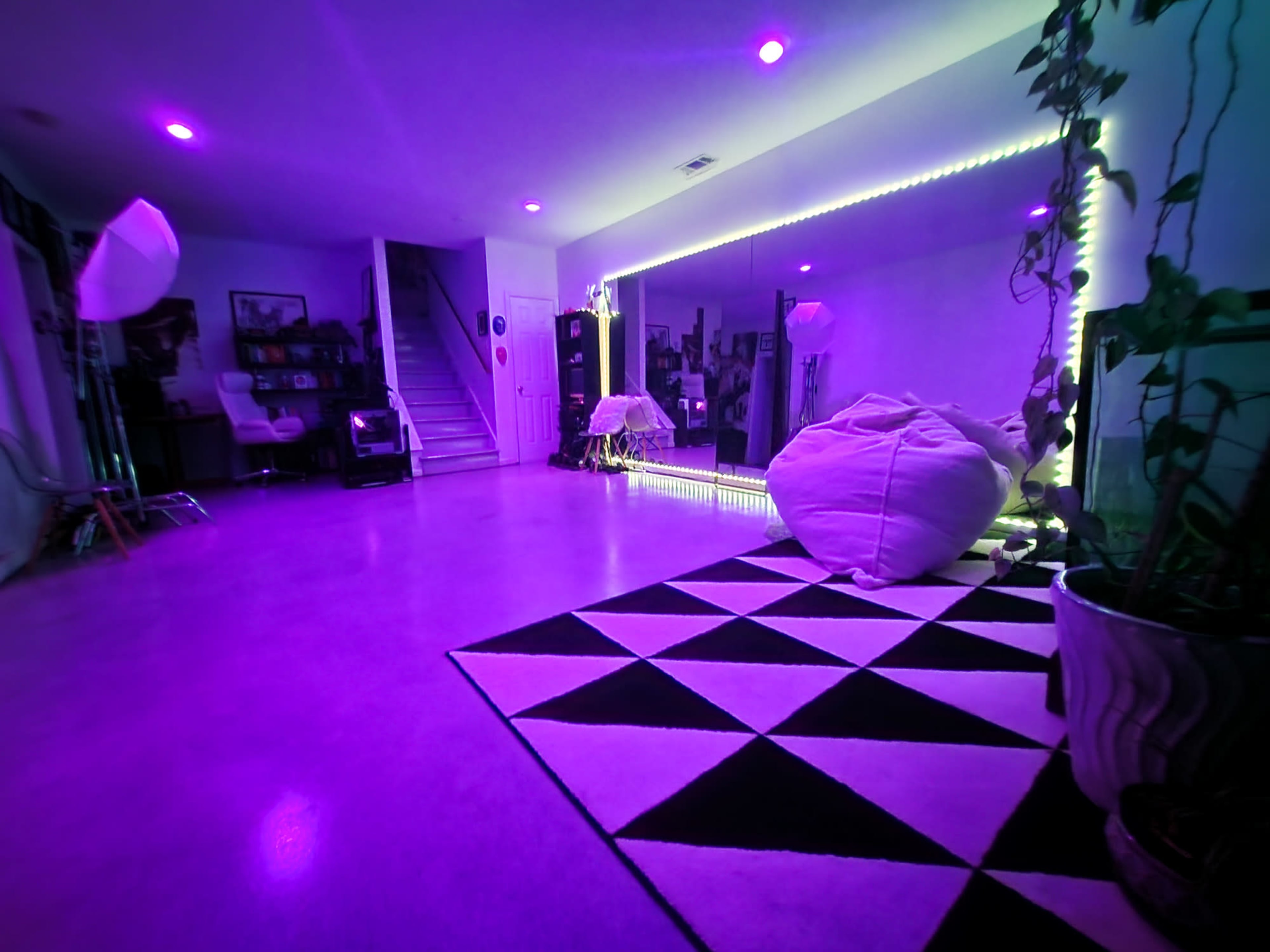 The image shows a dimly lit room with purple lighting, featuring a patterned rug, bean bags, mirrors, and decorative plants.