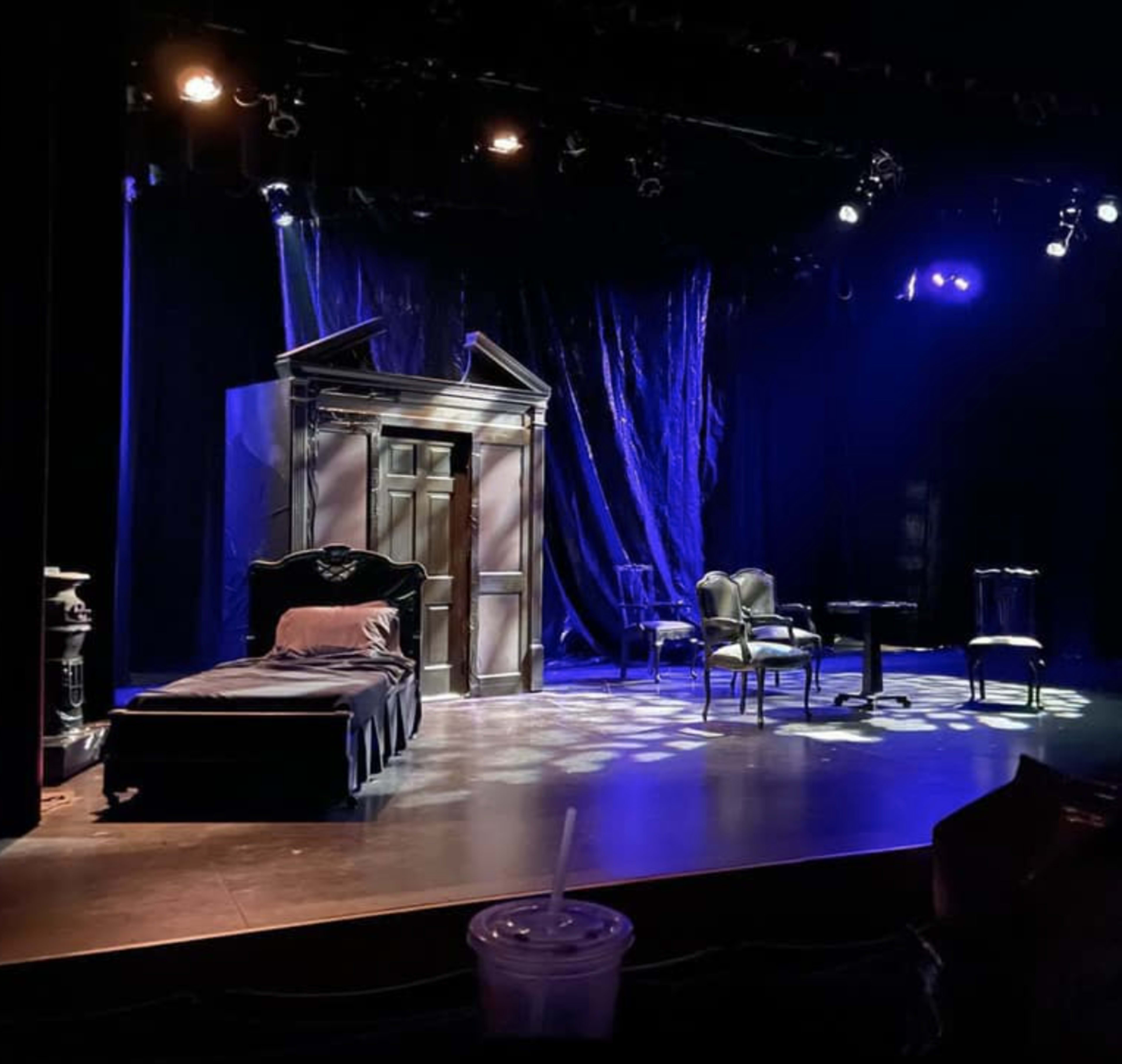 The image shows a dimly lit stage setup with a bed, an ornate chair, a small table, and a backdrop of dark fabric and lights.