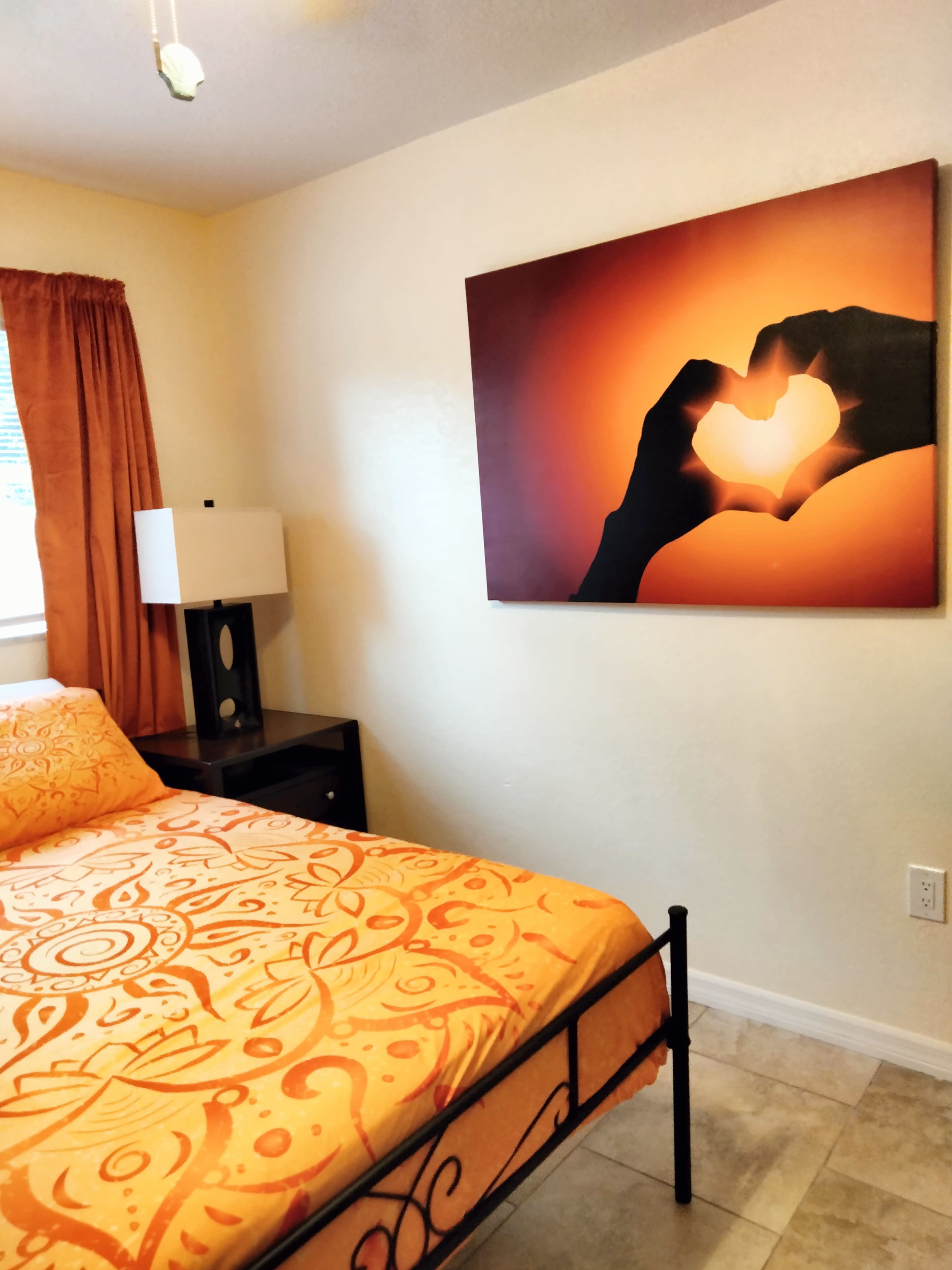 A bedroom features an orange bedspread, a nightstand with a lamp, and a wall art piece depicting two hands forming a heart.