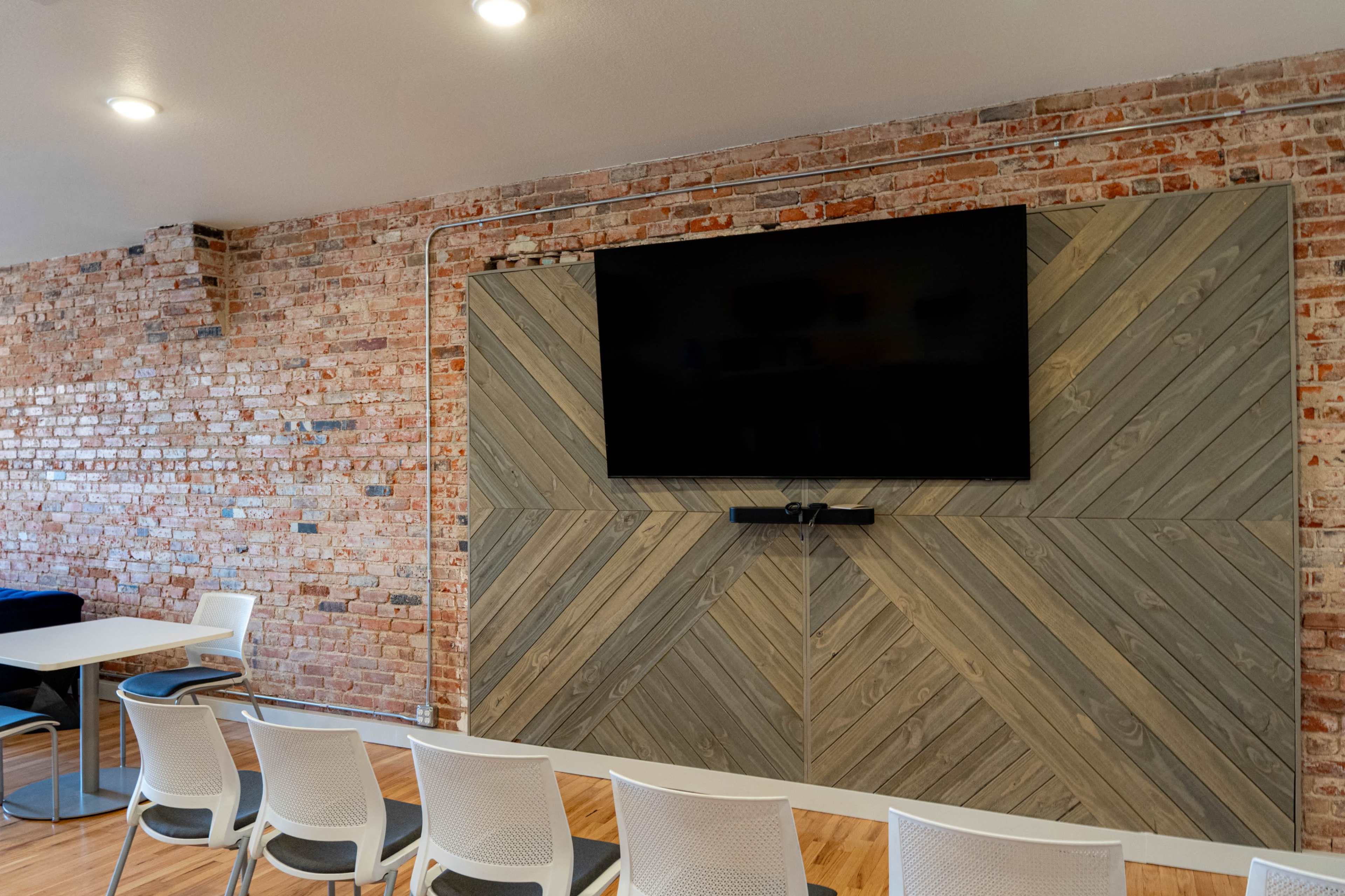 The image shows a meeting room with a large television mounted on a wooden wall, surrounded by several white chairs and a table.