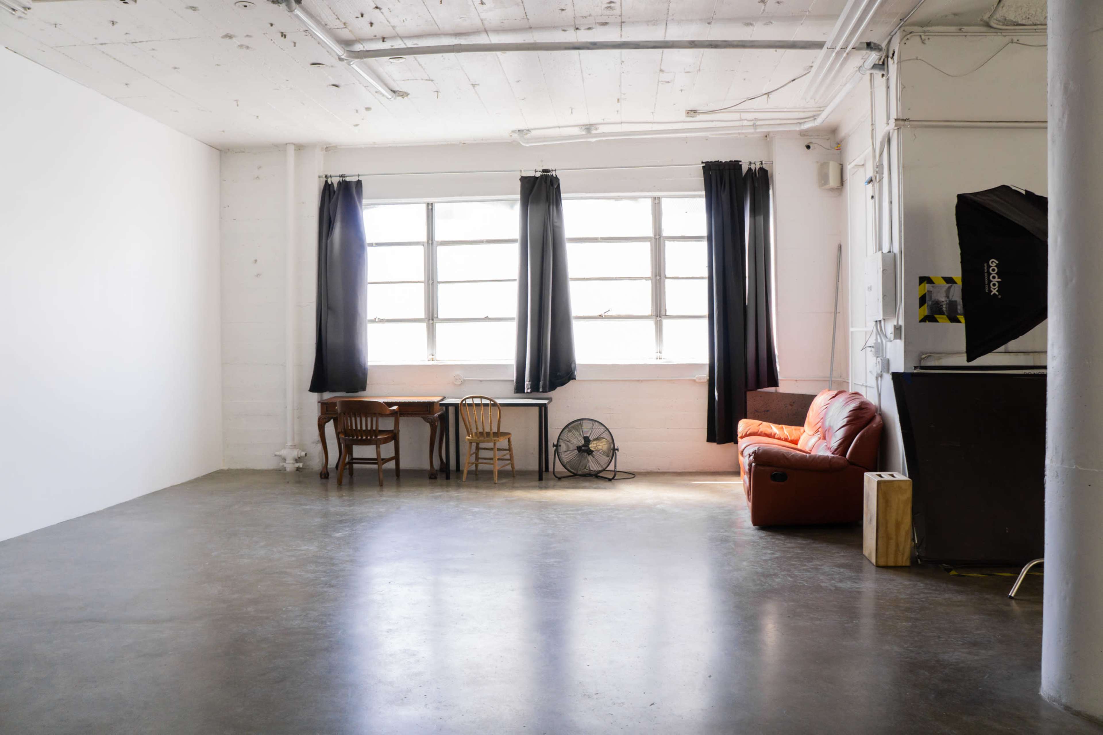 The image shows a spacious, minimalist room with two large windows, a brown leather sofa, a small wooden table with two chairs, a floor fan, and a backdrop stand in the corner.