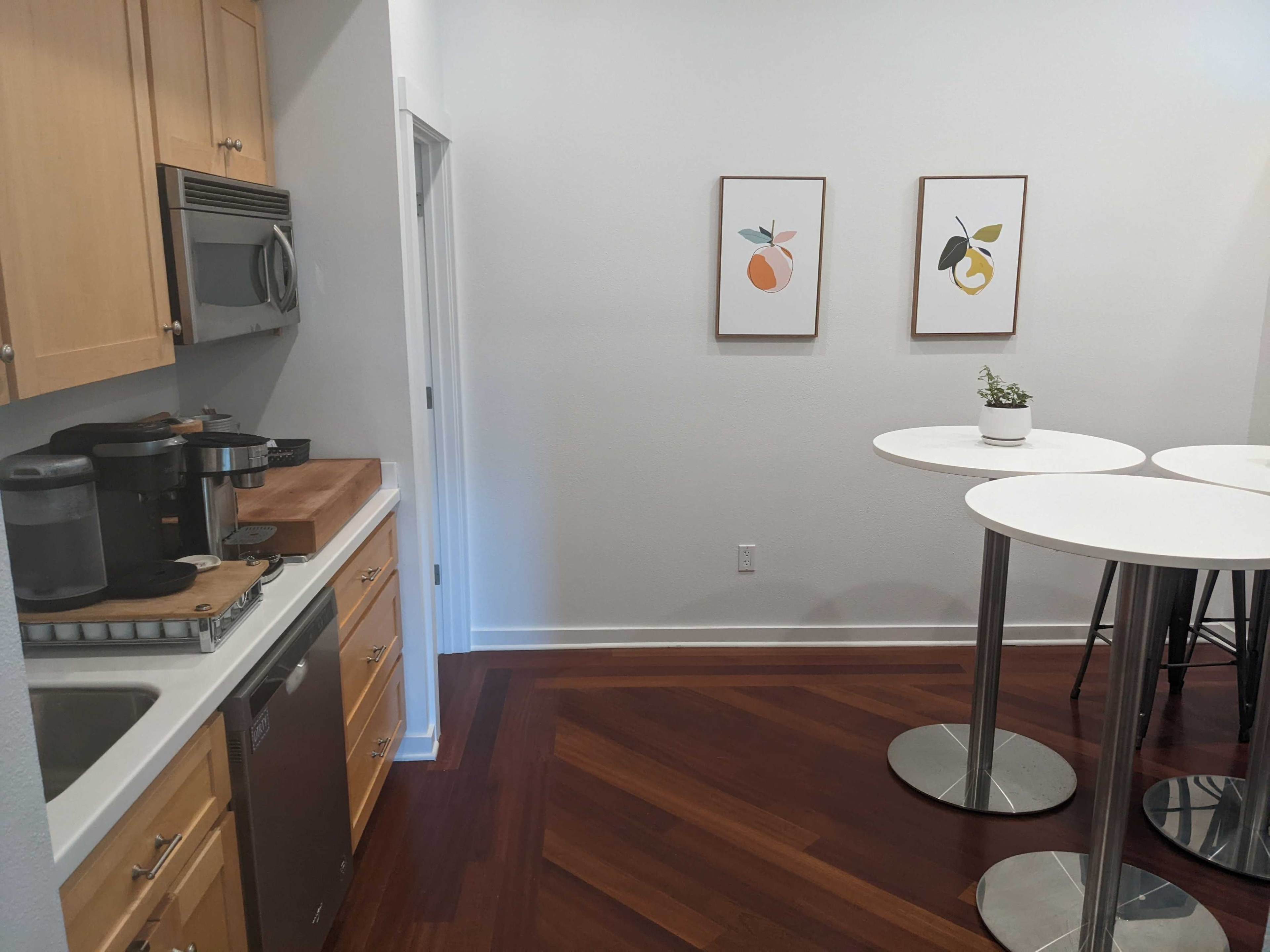 A modern kitchen area with wooden cabinets, a stainless steel sink, and two round white tables with metallic bases, adorned with framed fruit prints on the wall.