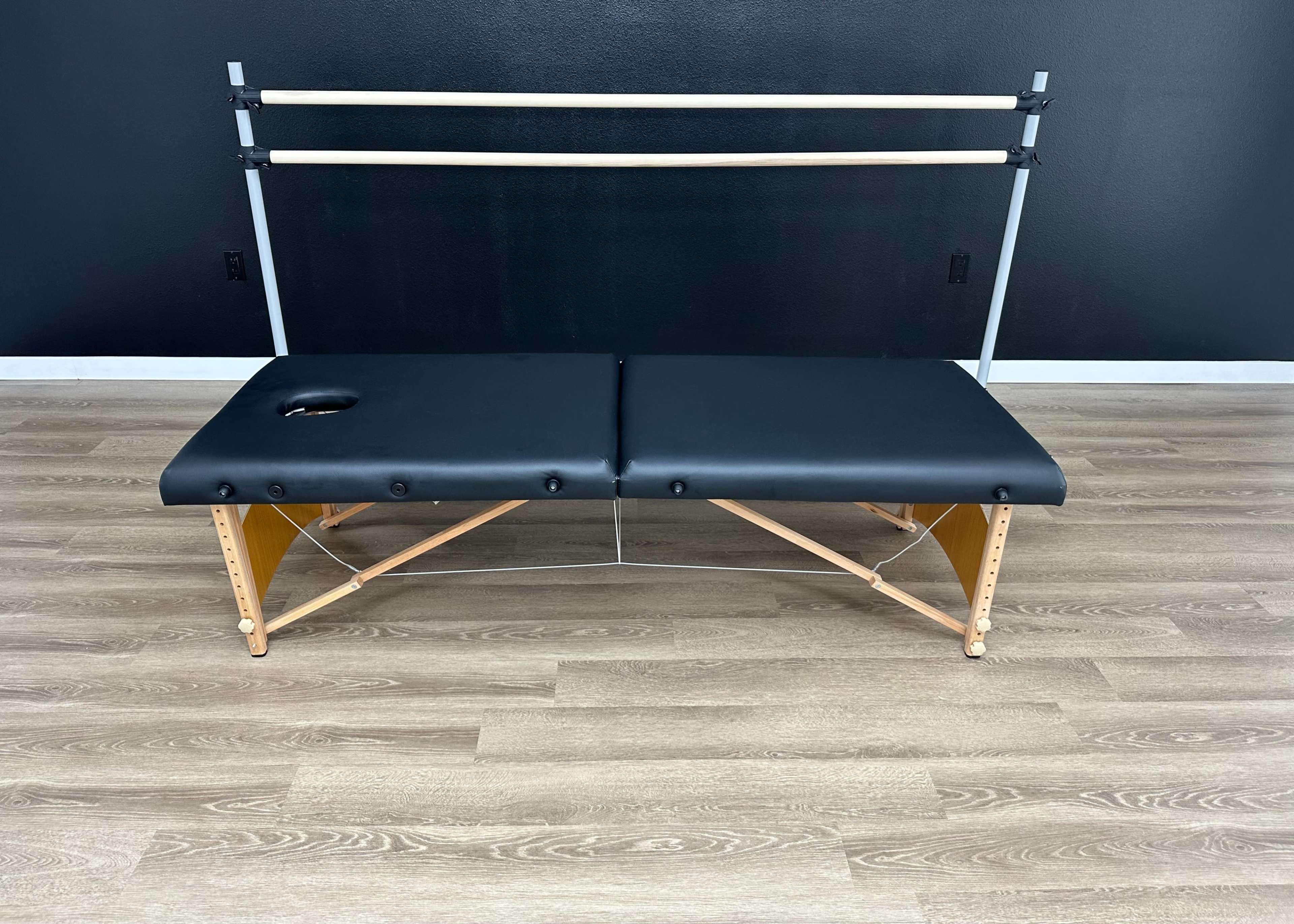A black massage table with a wooden frame, set against a dark wall and a simple bar mounted above it.