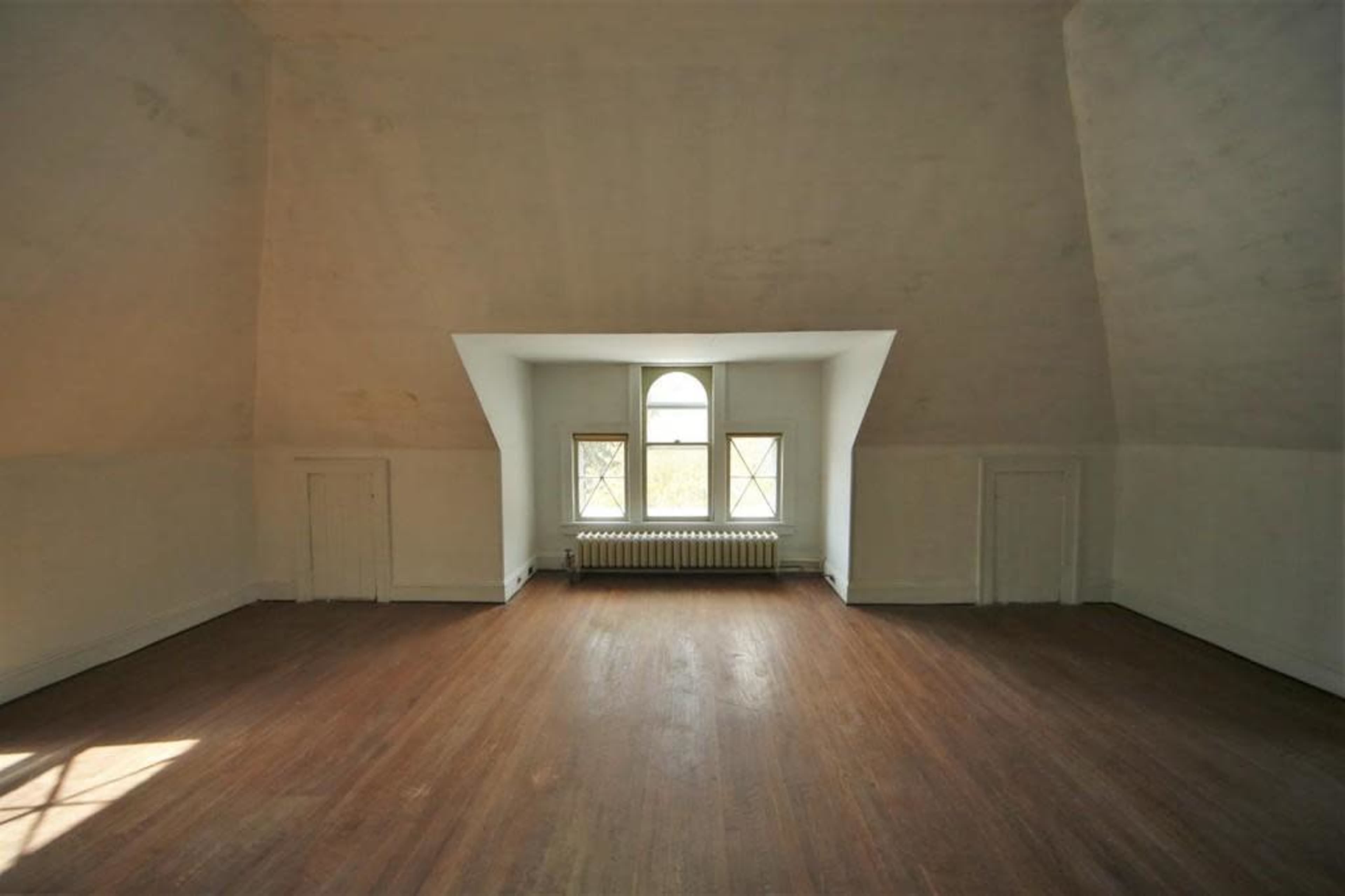 An empty room with a sloped ceiling features a large window and a wooden floor.