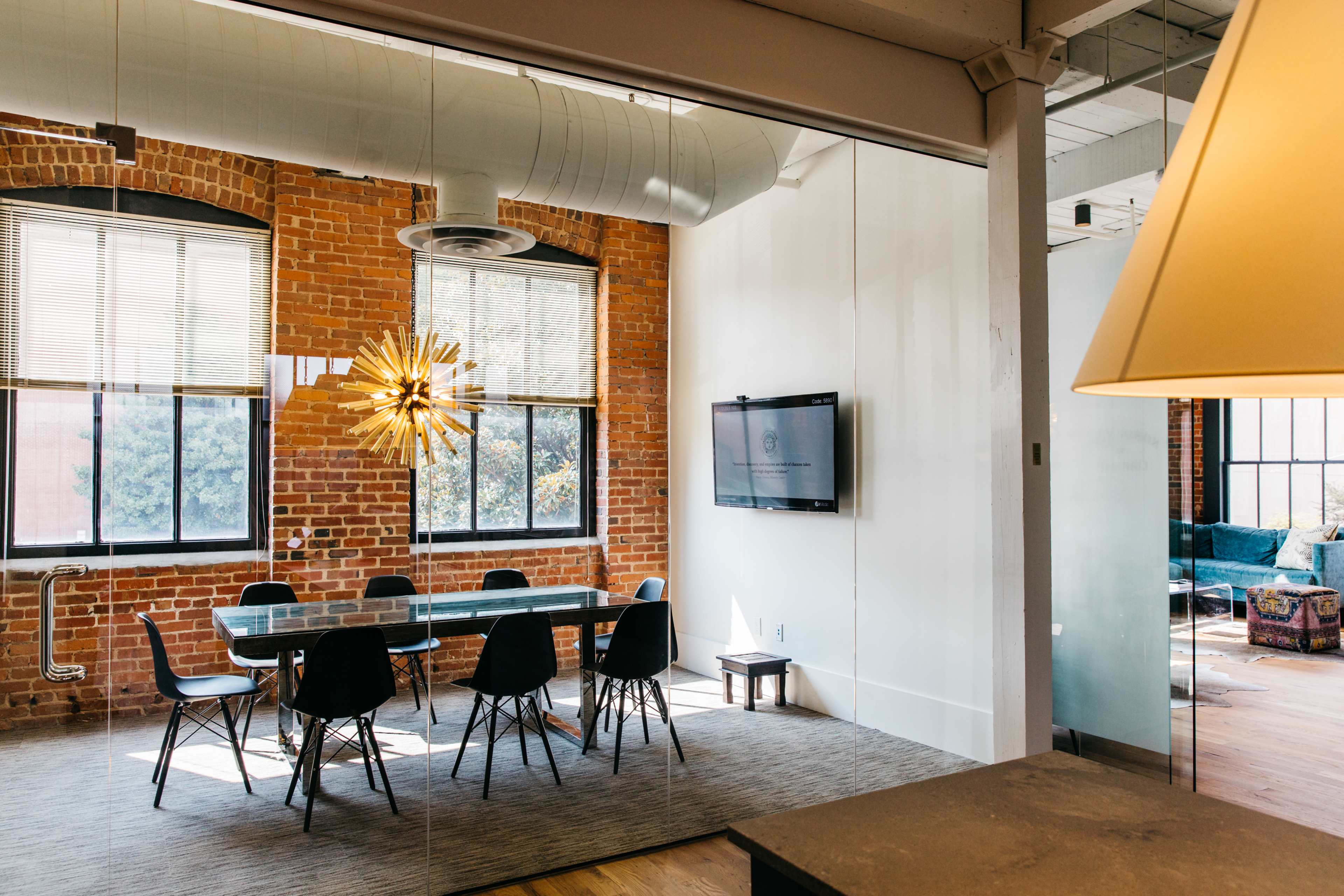 A bright conference room features a long table surrounded by black chairs, with large windows exposing brick walls and a television mounted on the wall.