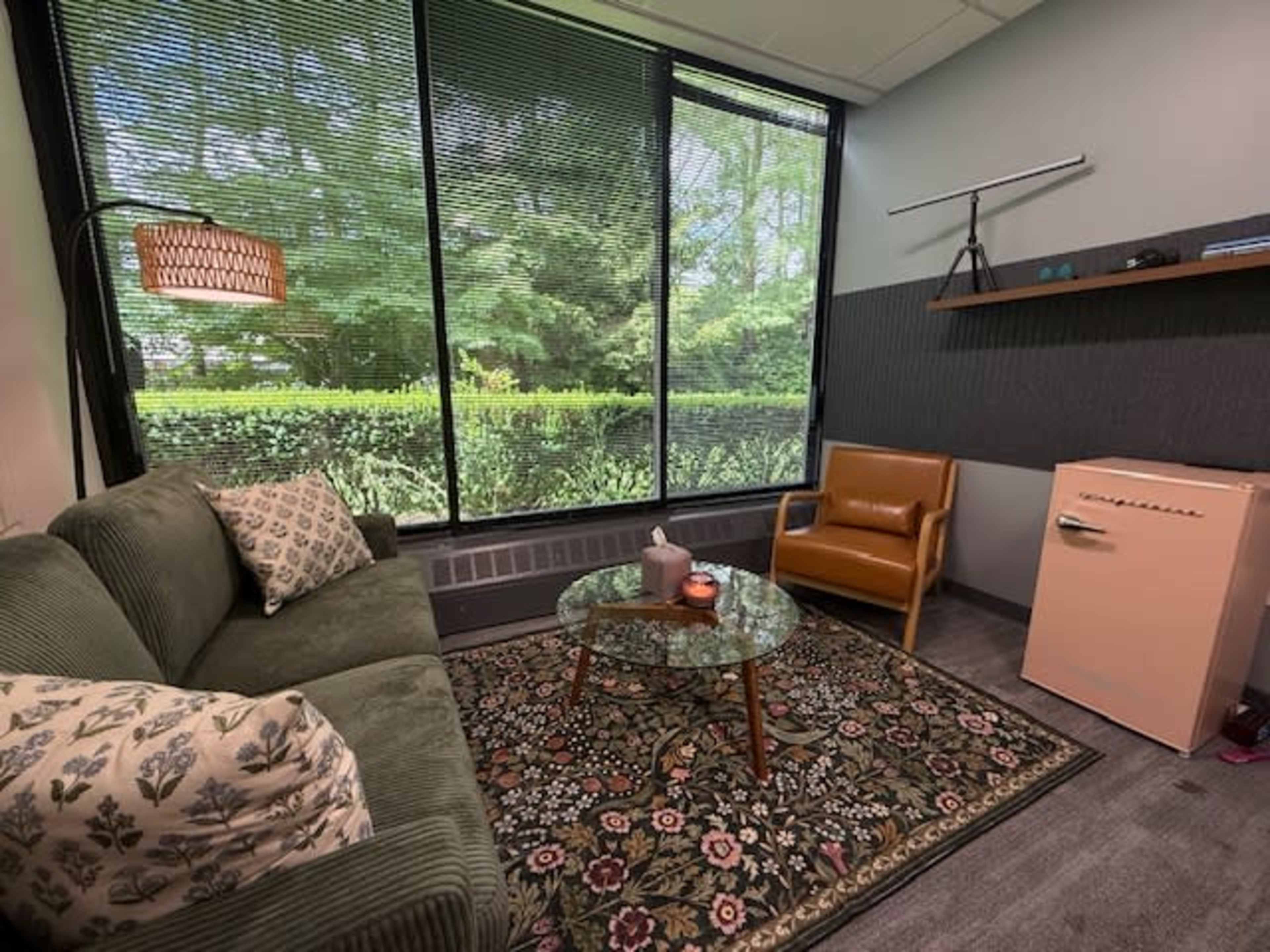 The image shows a small, well-lit room with a green couch, a brown armchair, a glass coffee table on a patterned rug, and a vintage-style refrigerator against the wall, surrounded by large windows with views of greenery outside.