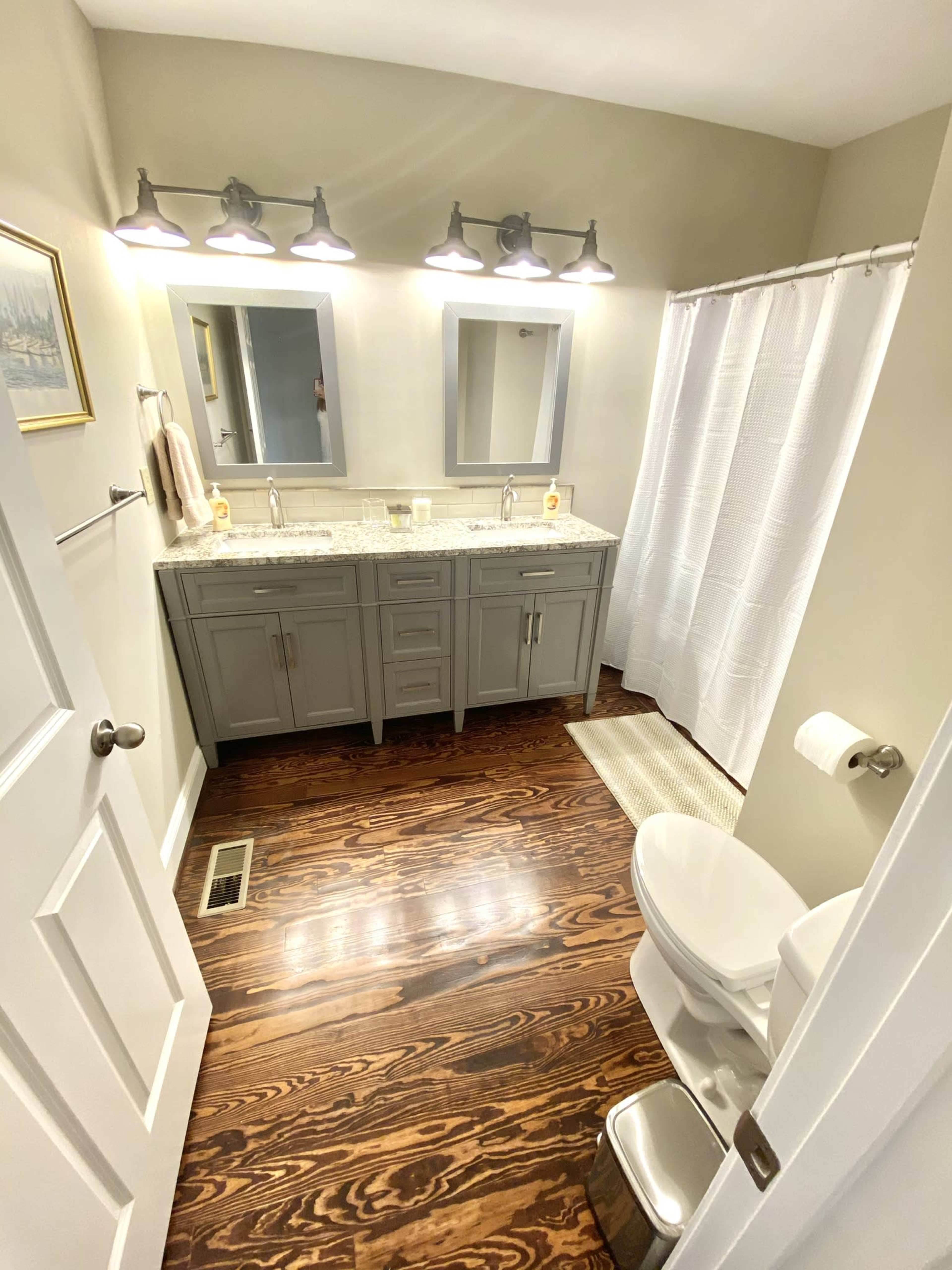 A modern bathroom with a double vanity, two mirrors, a shower curtain, and wooden flooring.