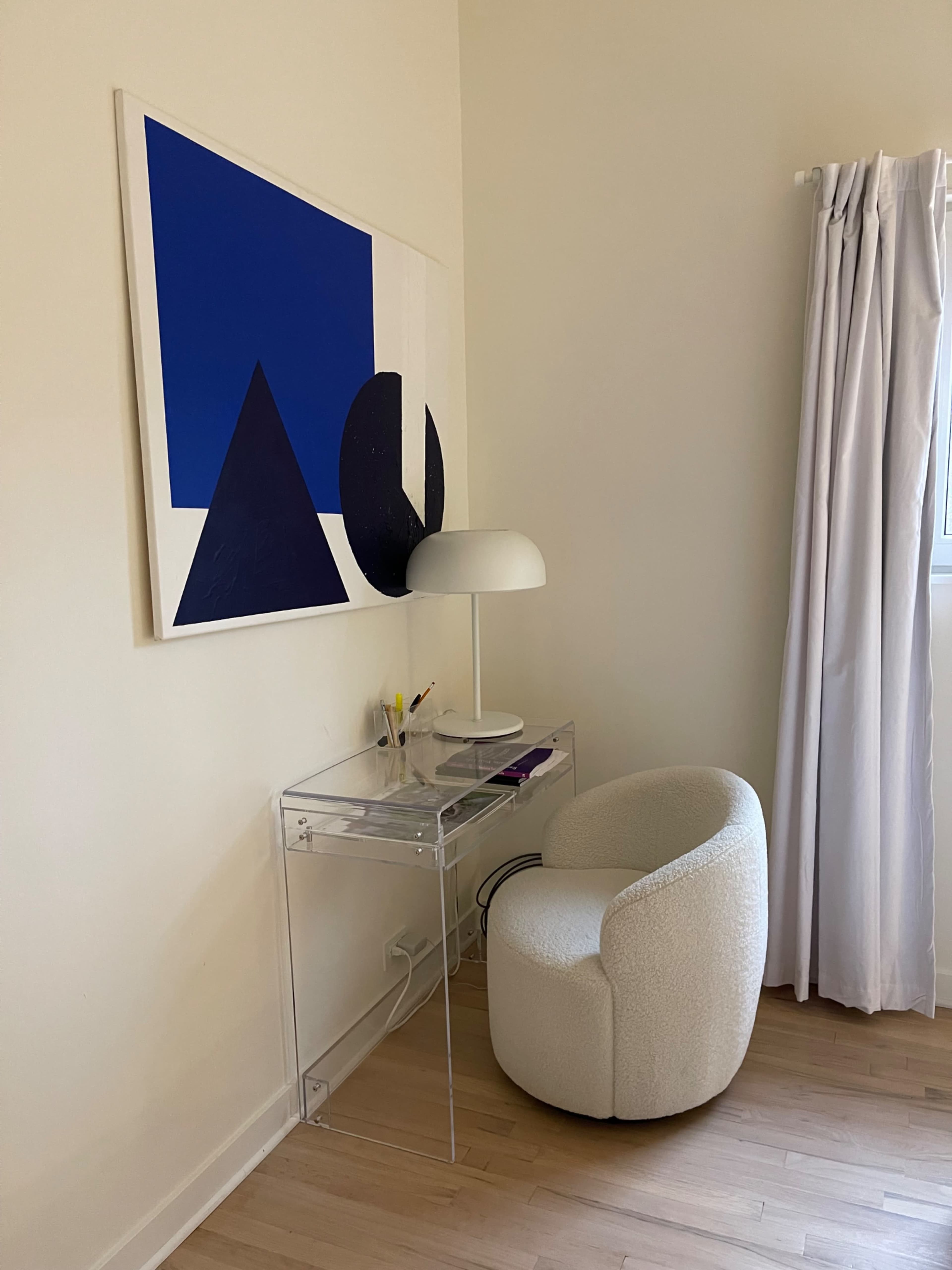 A clear acrylic desk with a white lamp and art piece hangs on the wall beside a round, upholstered chair.