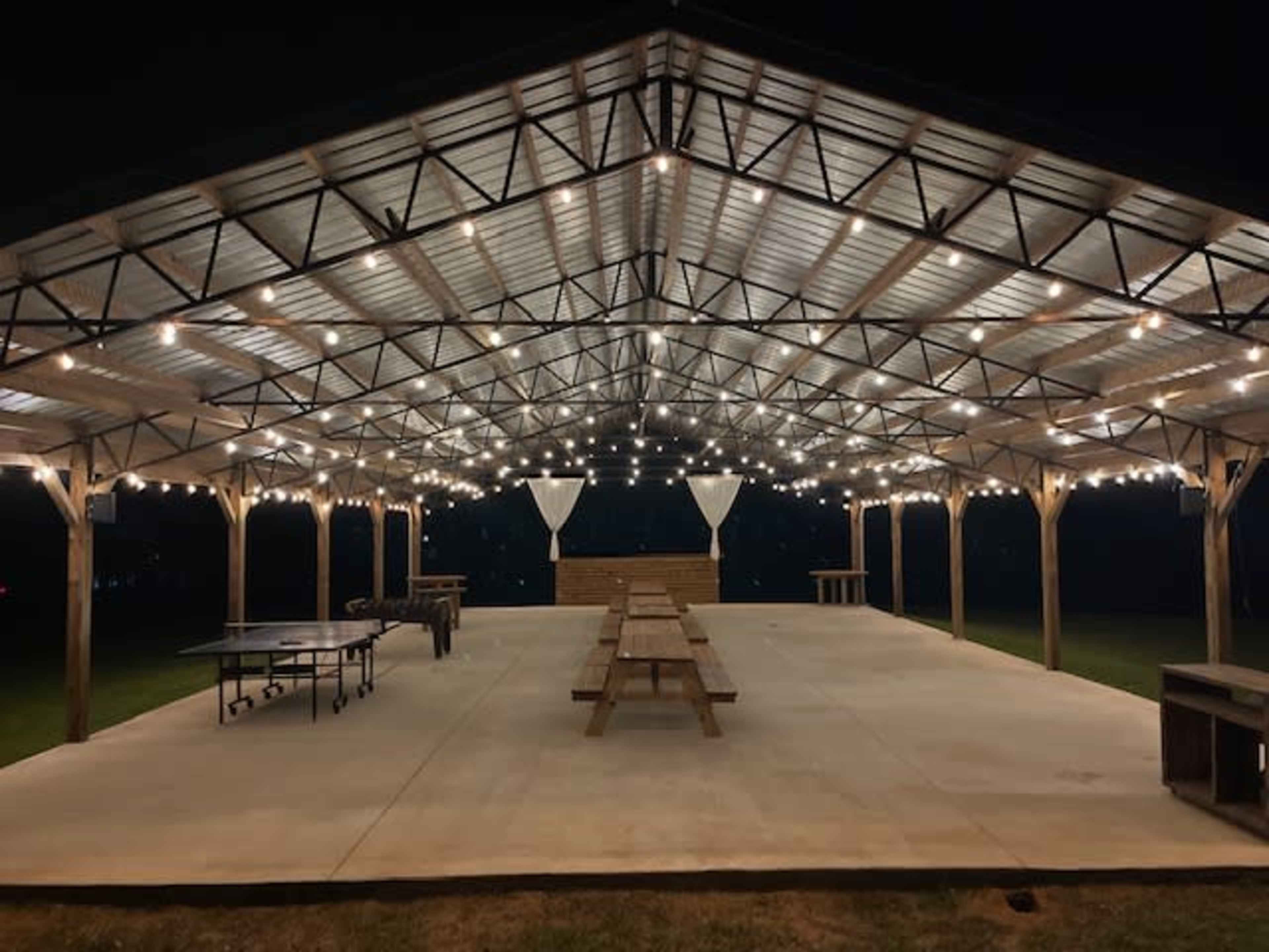 A large, open pavilion with string lights hanging from the ceiling, featuring multiple picnic tables and a concrete floor, set in a grassy area at night.