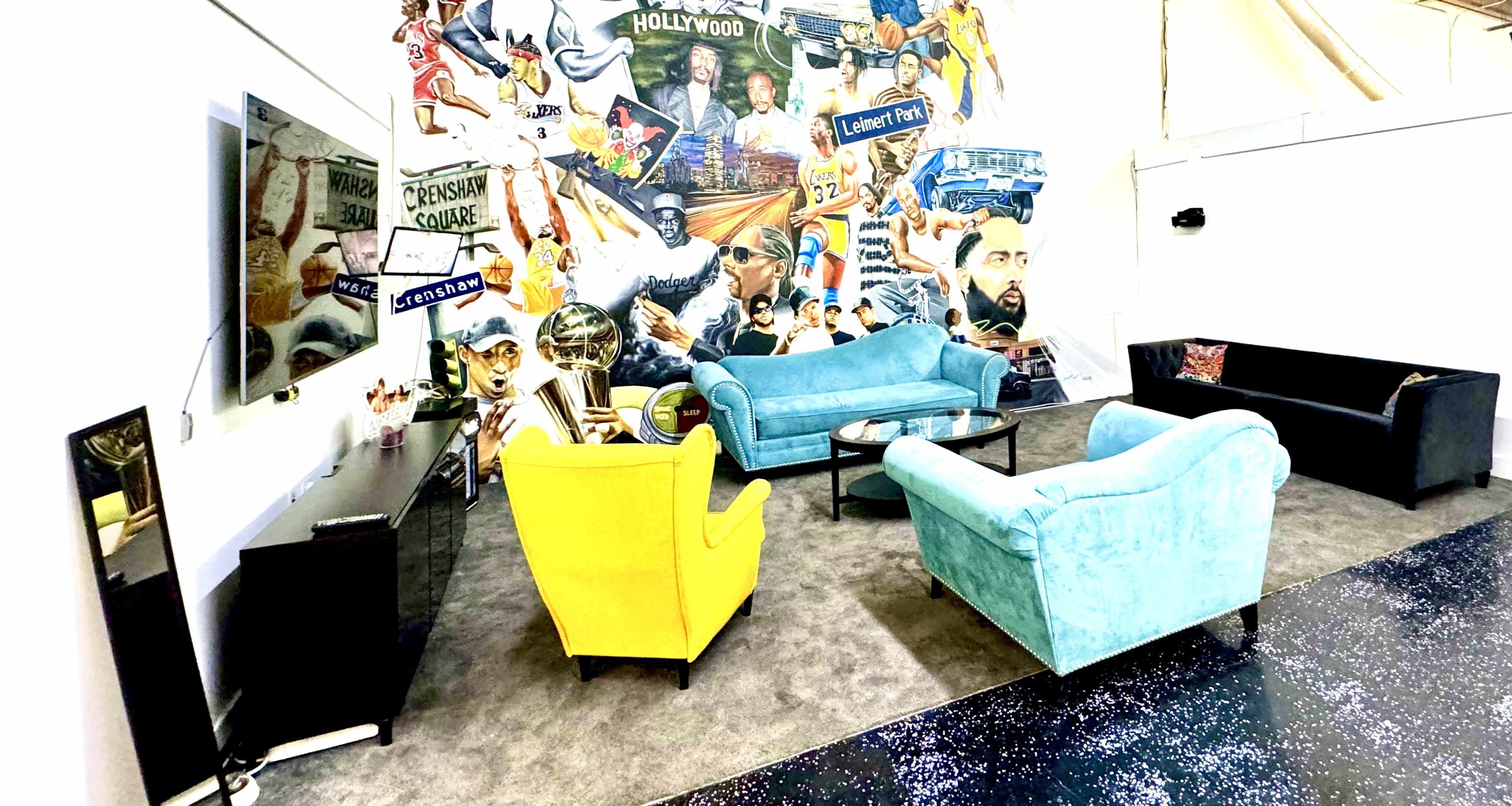 A modern lounge area features two sofas and two armchairs arranged around a coffee table, with a vibrant mural depicting various cultural icons on the wall behind.