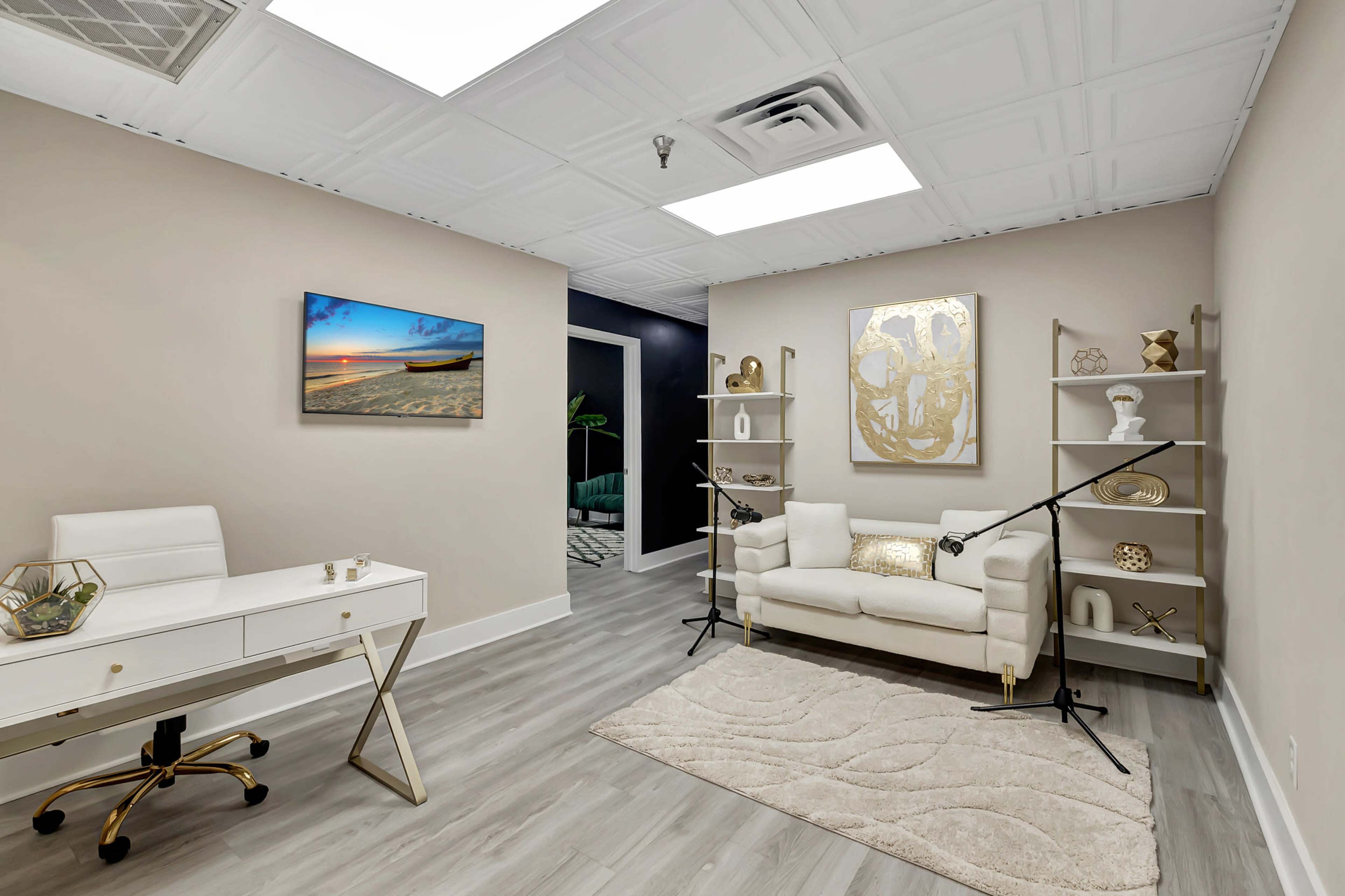 A modern office space featuring a white desk with a chair, a light-colored couch, decorative shelves, and a beach scene painting on the wall.
