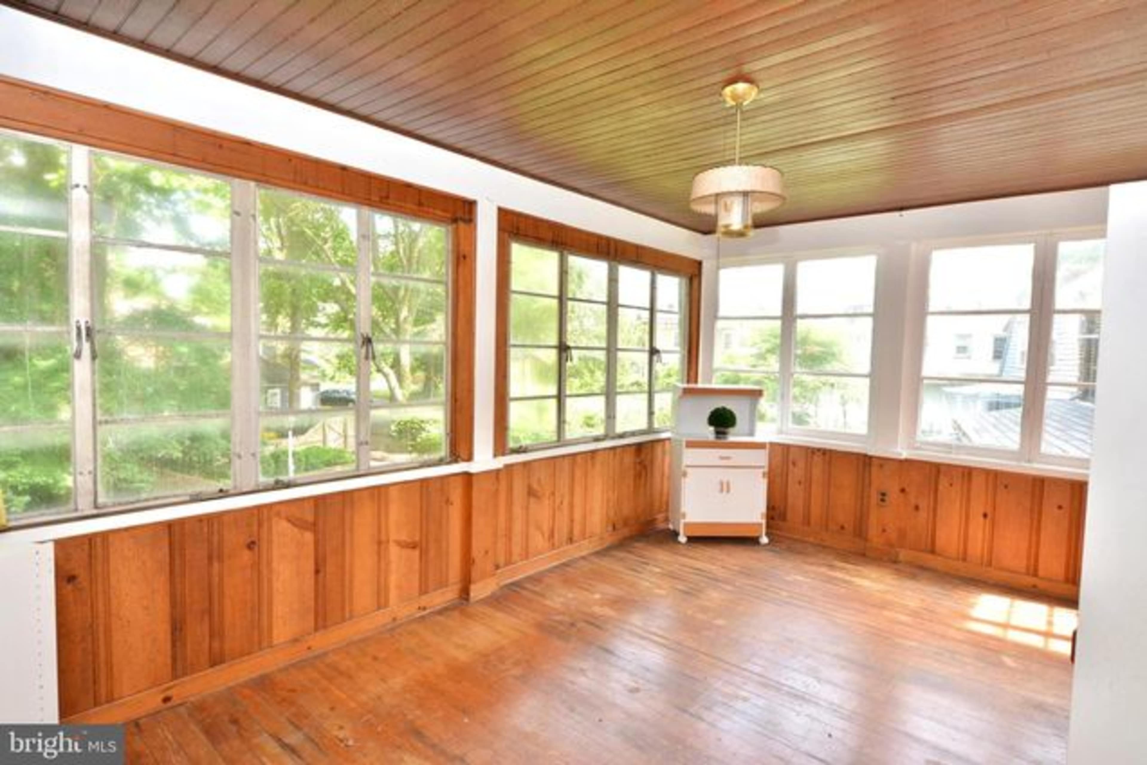 A sunlit room with wooden walls, a wooden floor, and large windows allowing natural light to enter.