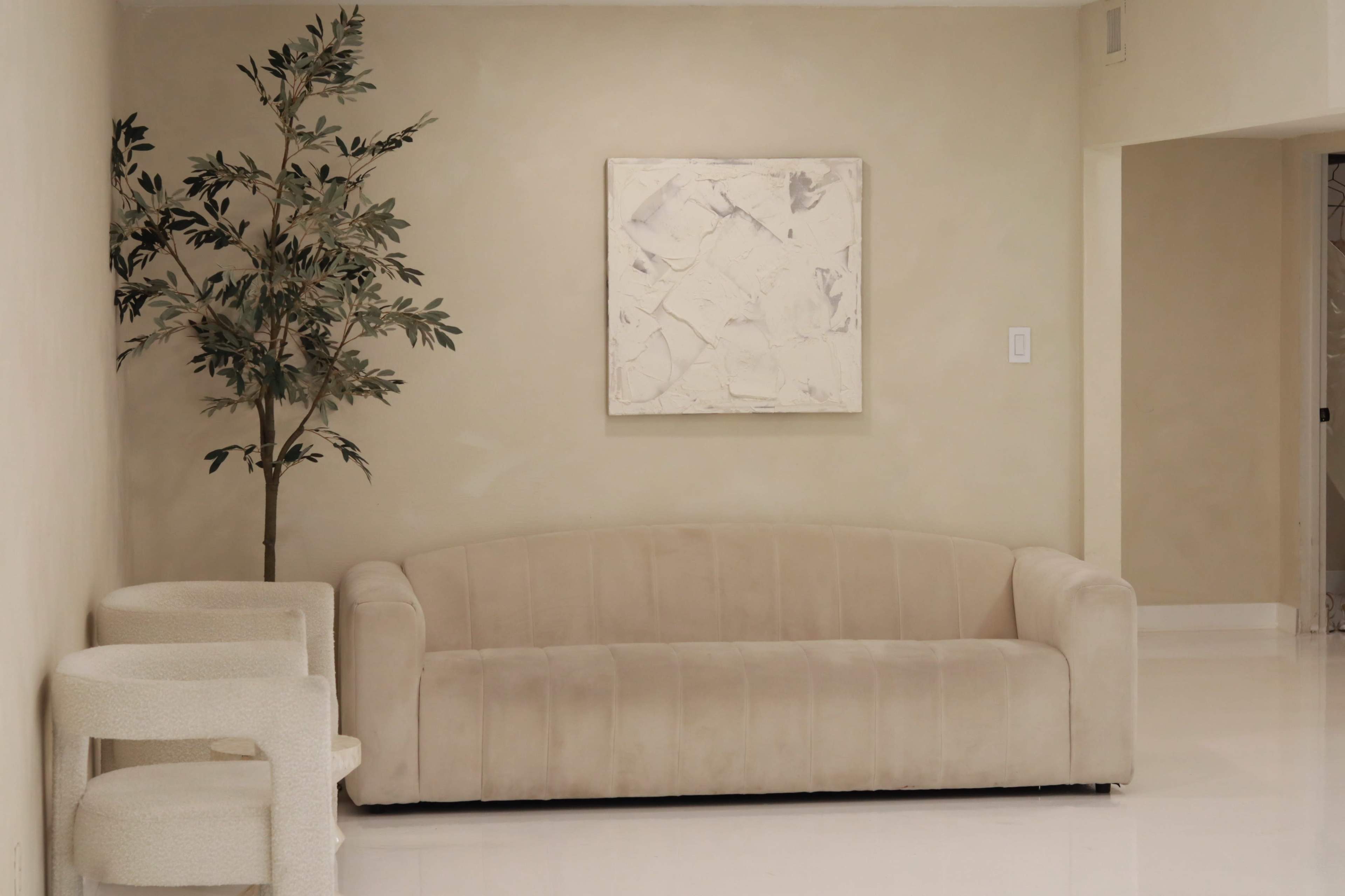 A beige couch is positioned against a light-colored wall next to a small potted tree and a piece of abstract art.