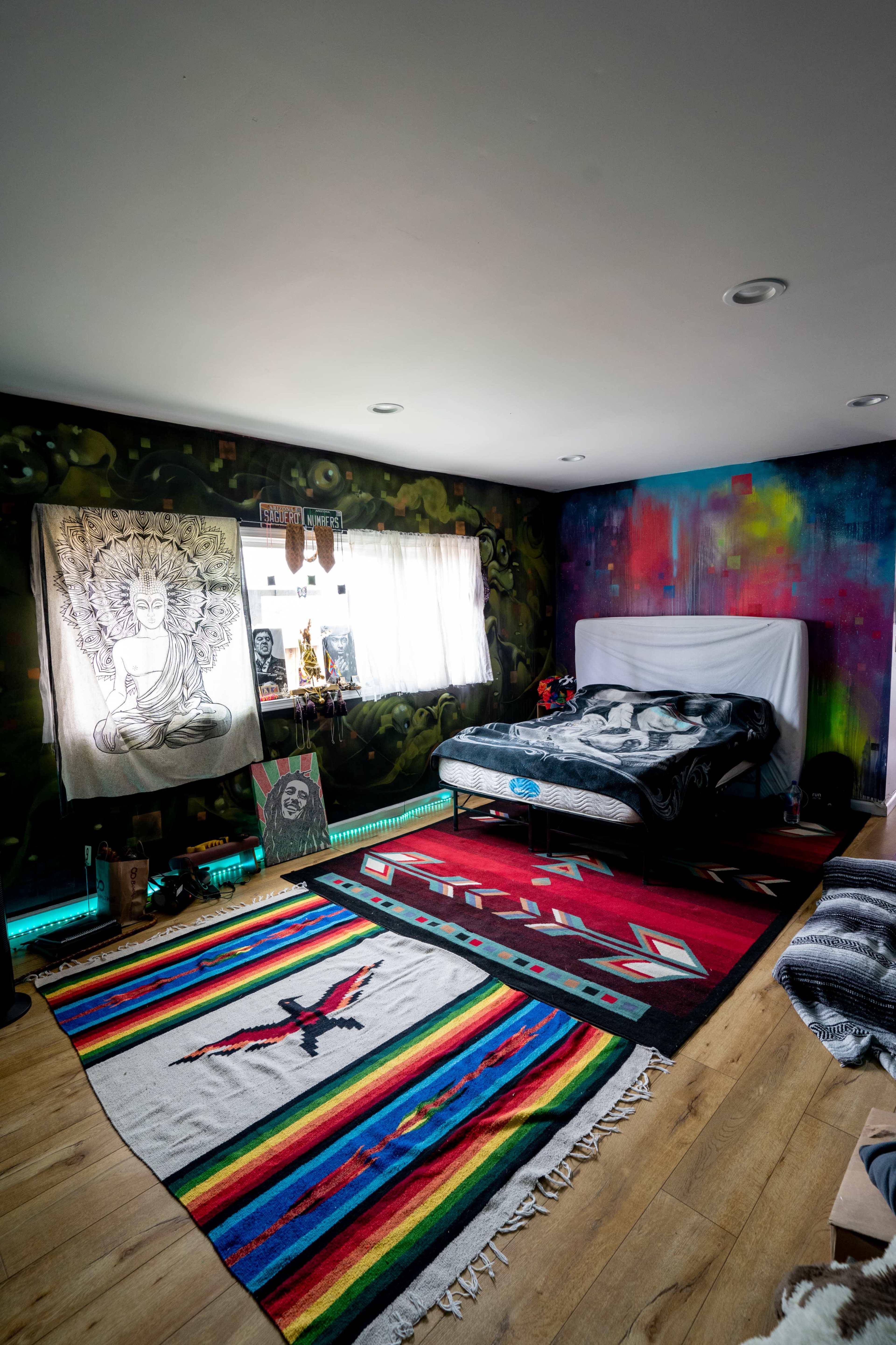 A minimalist bedroom features a bed against a wall adorned with decorative fabric, colorful rugs on a wooden floor, and various artworks displayed on the walls.