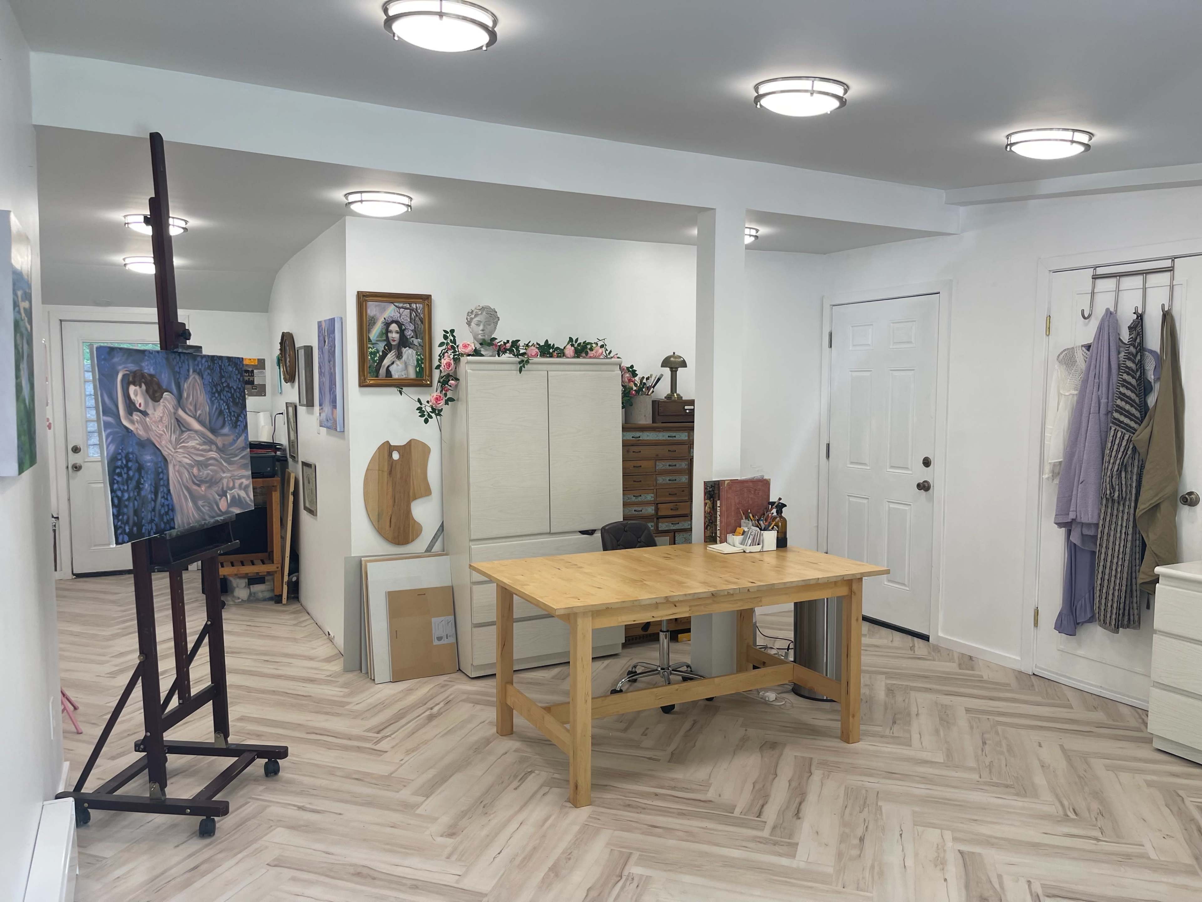 The image shows a well-lit art studio featuring a wooden table, an easel with a painting, storage cabinets, and artwork displayed on the walls.