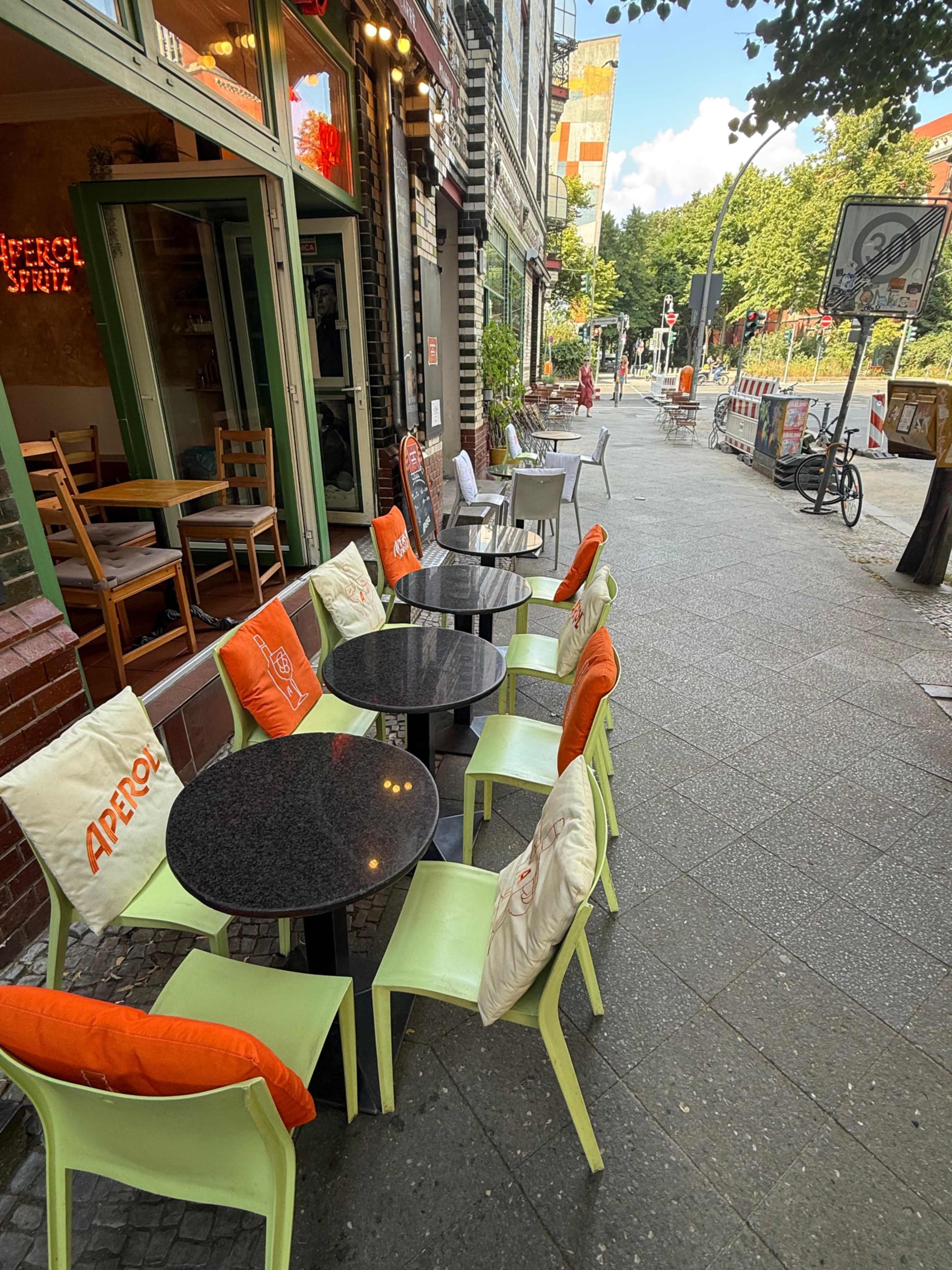 A row of tables with chairs, some adorned with cushions, is lined along a sidewalk outside a café, with a vibrant urban setting in the background.