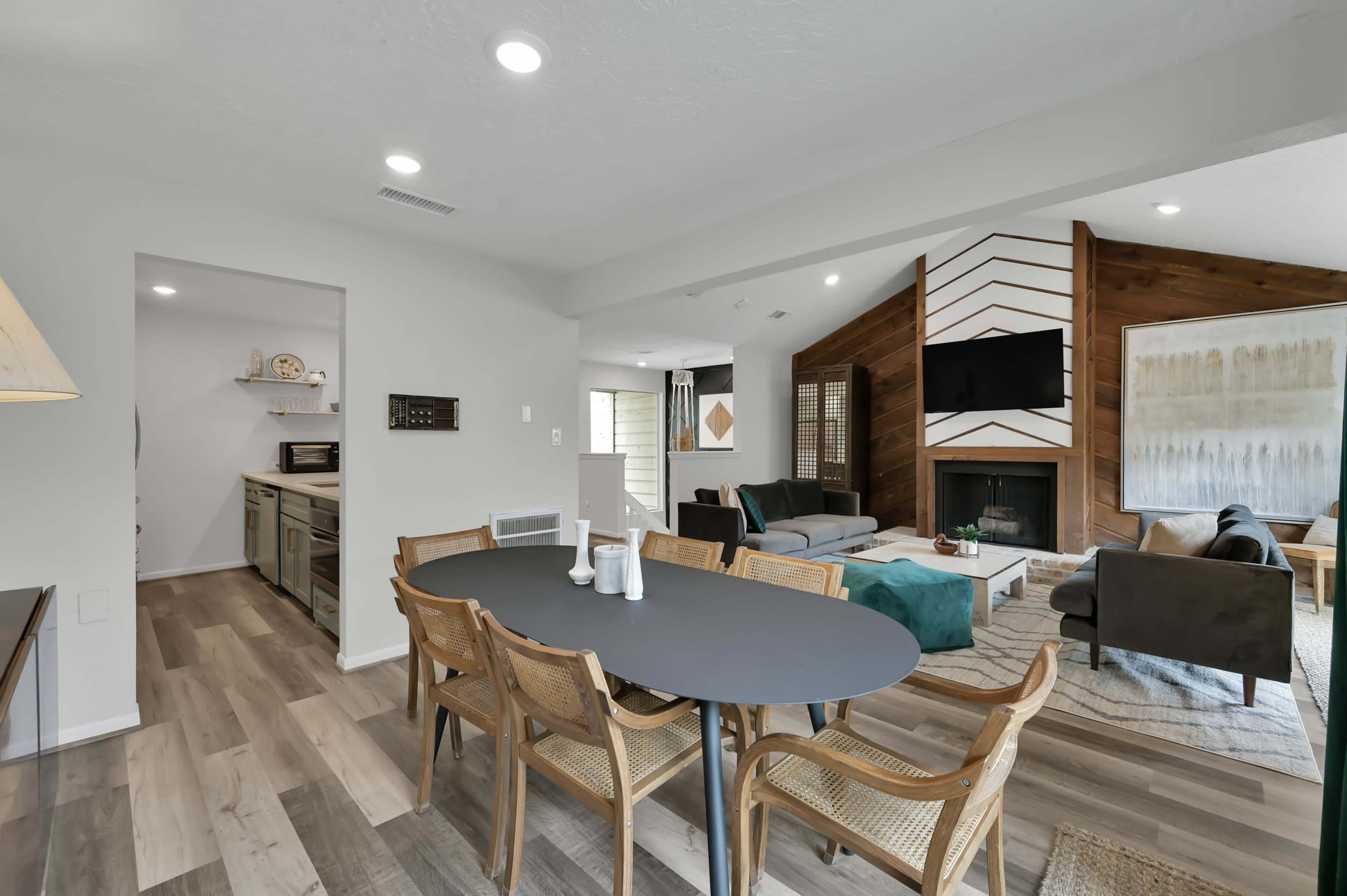 The image shows a modern living space featuring a dining area with a dark table and woven chairs, connected to a cozy living room with a fireplace and a wall-mounted TV.