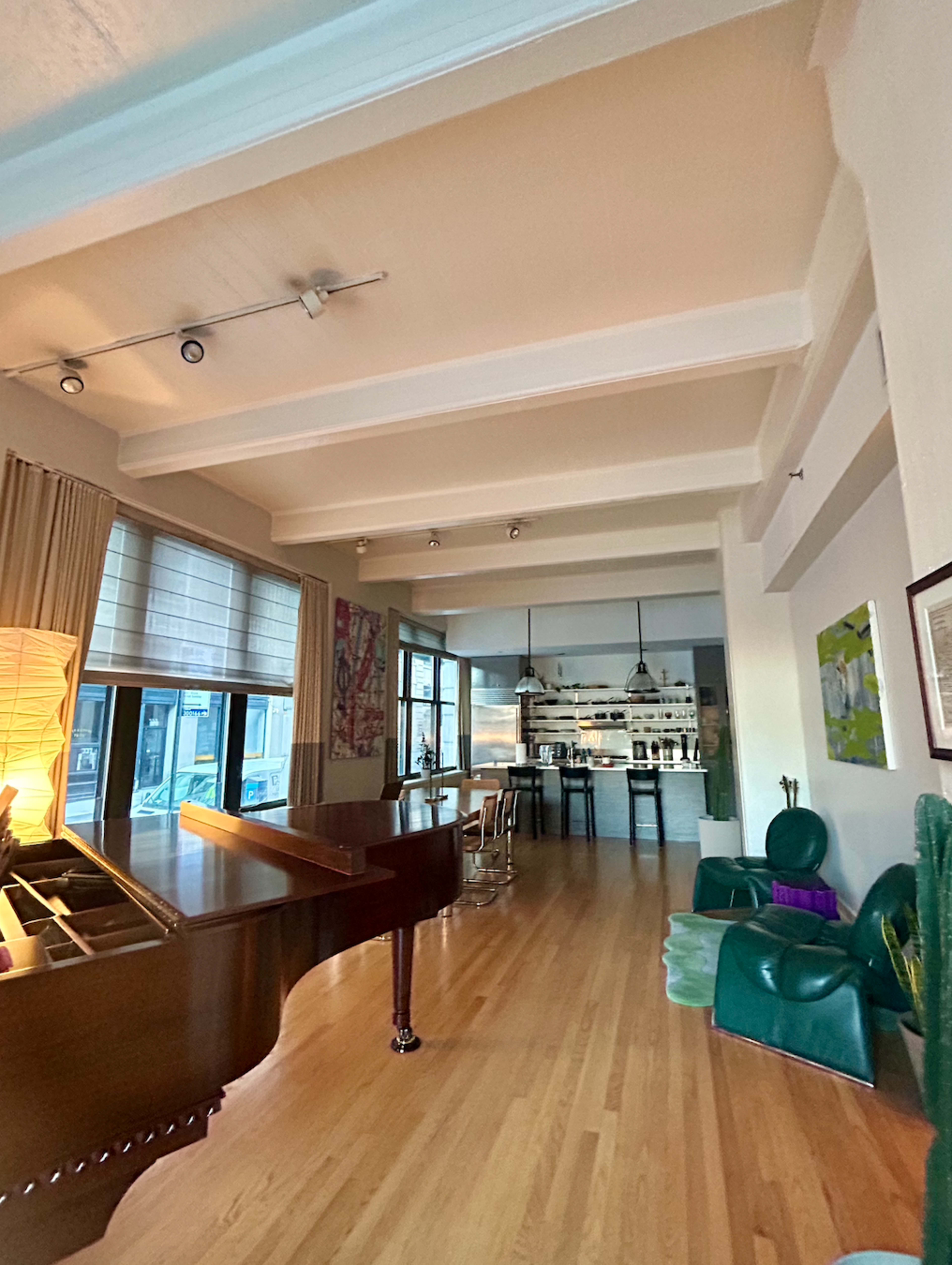 A spacious room features a grand piano, large windows, a kitchen area in the background, and contemporary furniture.