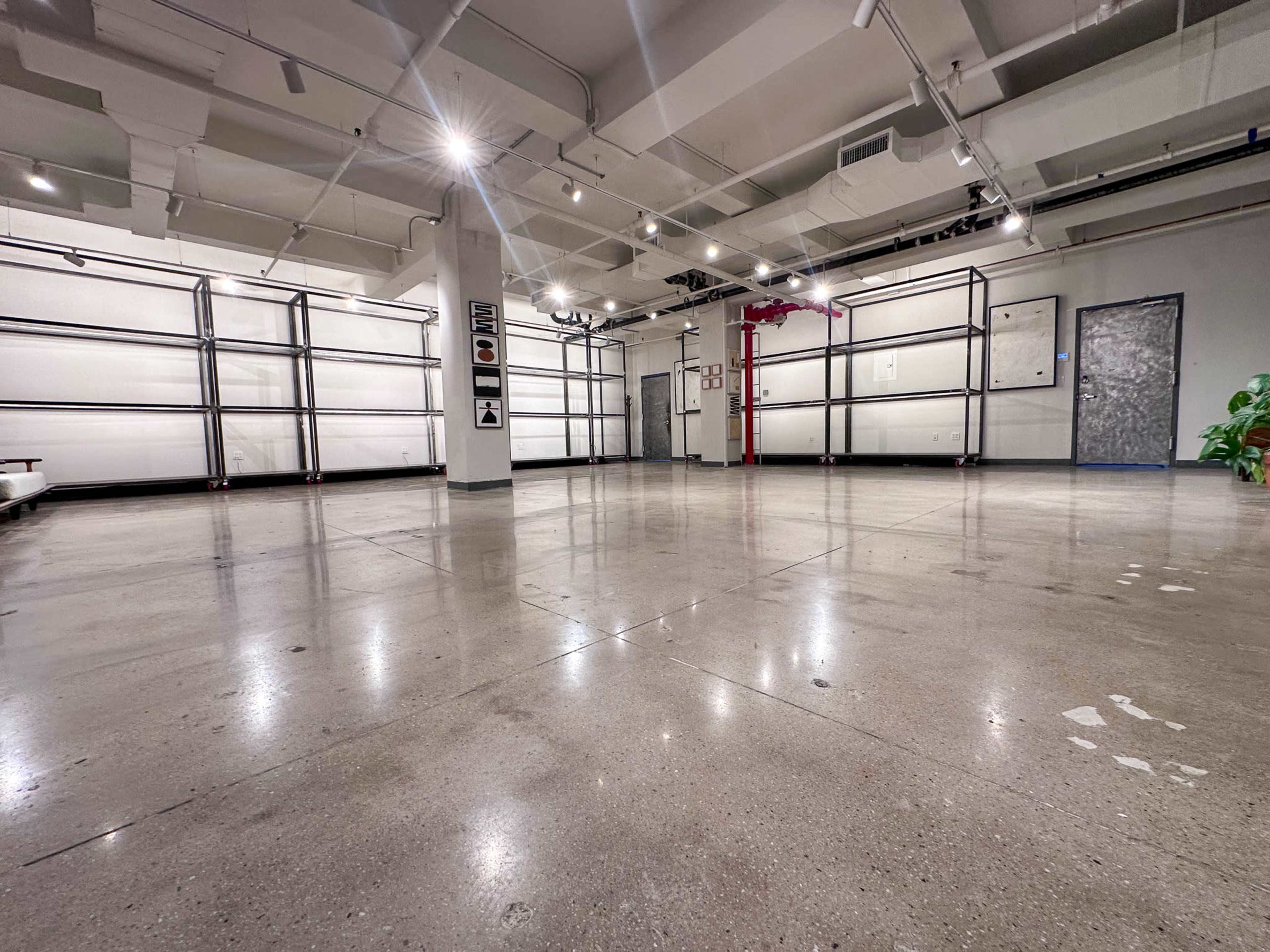 The image shows a spacious, minimally furnished interior with concrete flooring and metal framework along the walls.