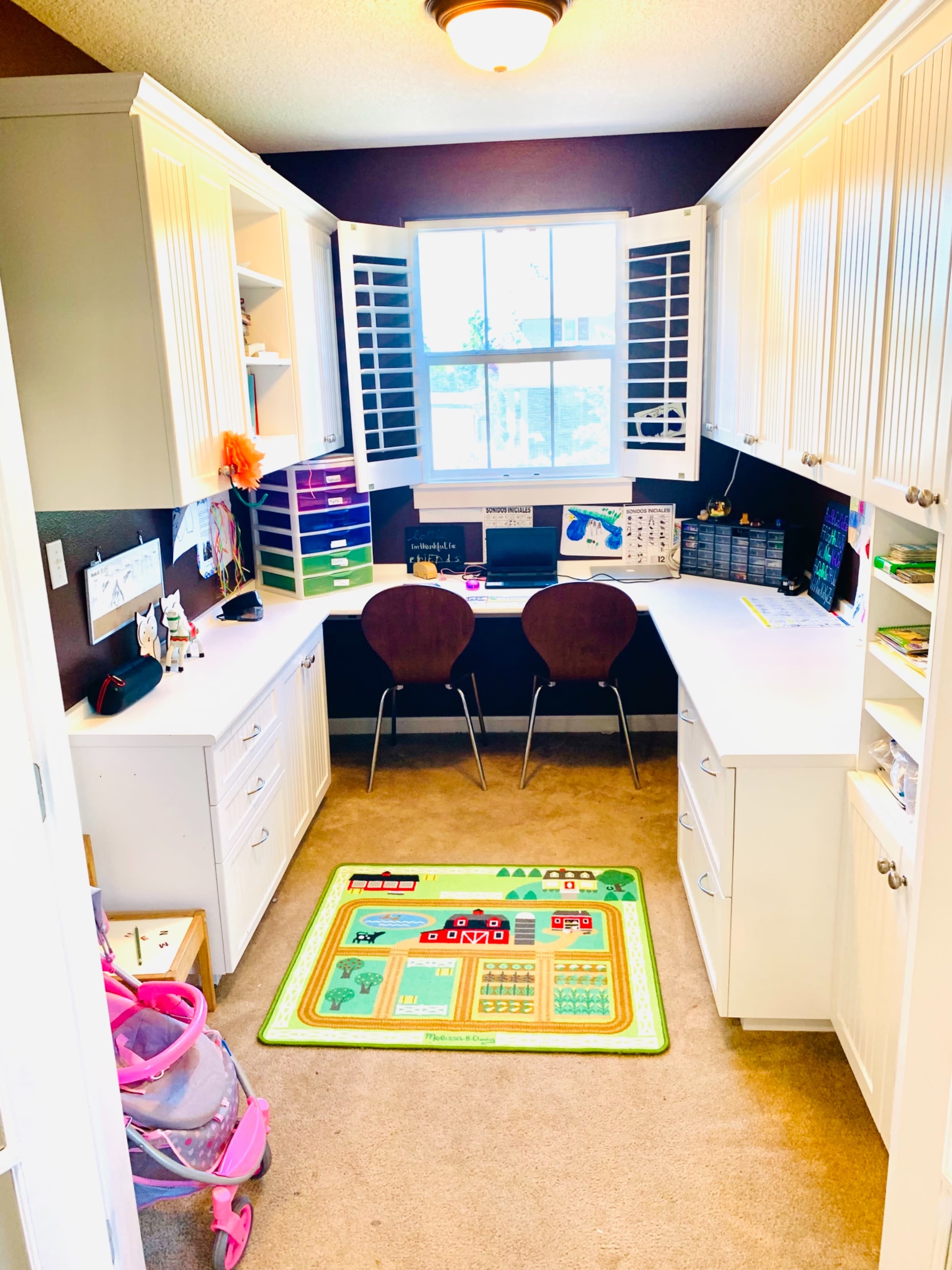 A home office features a corner desk setup with two chairs, storage cabinets, and a play mat on the carpeted floor.
