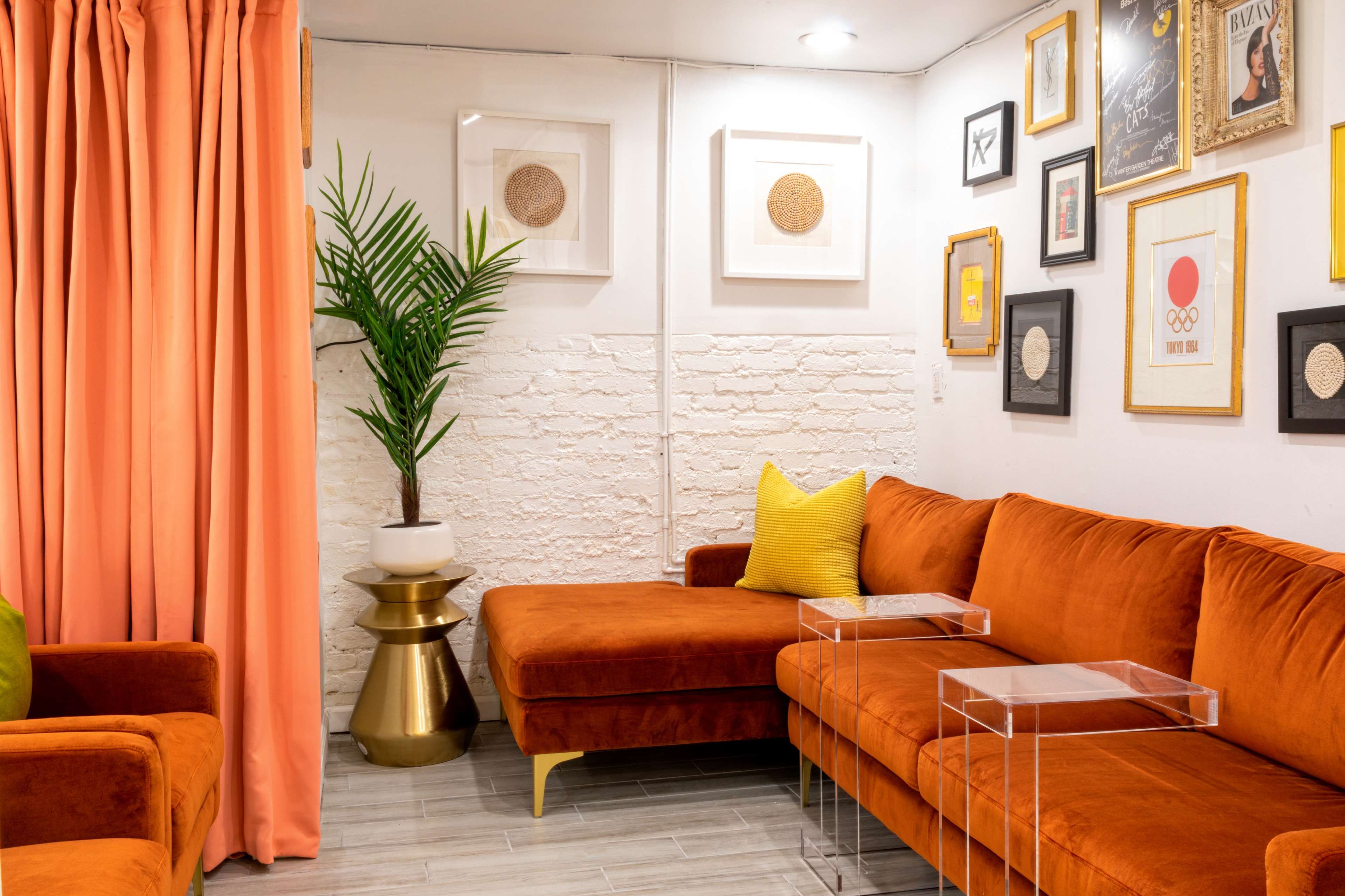 A cozy seating area with an orange sectional sofa, a small glass side table, a gold accent table, and framed artwork on the walls, complemented by a potted plant and curtains.