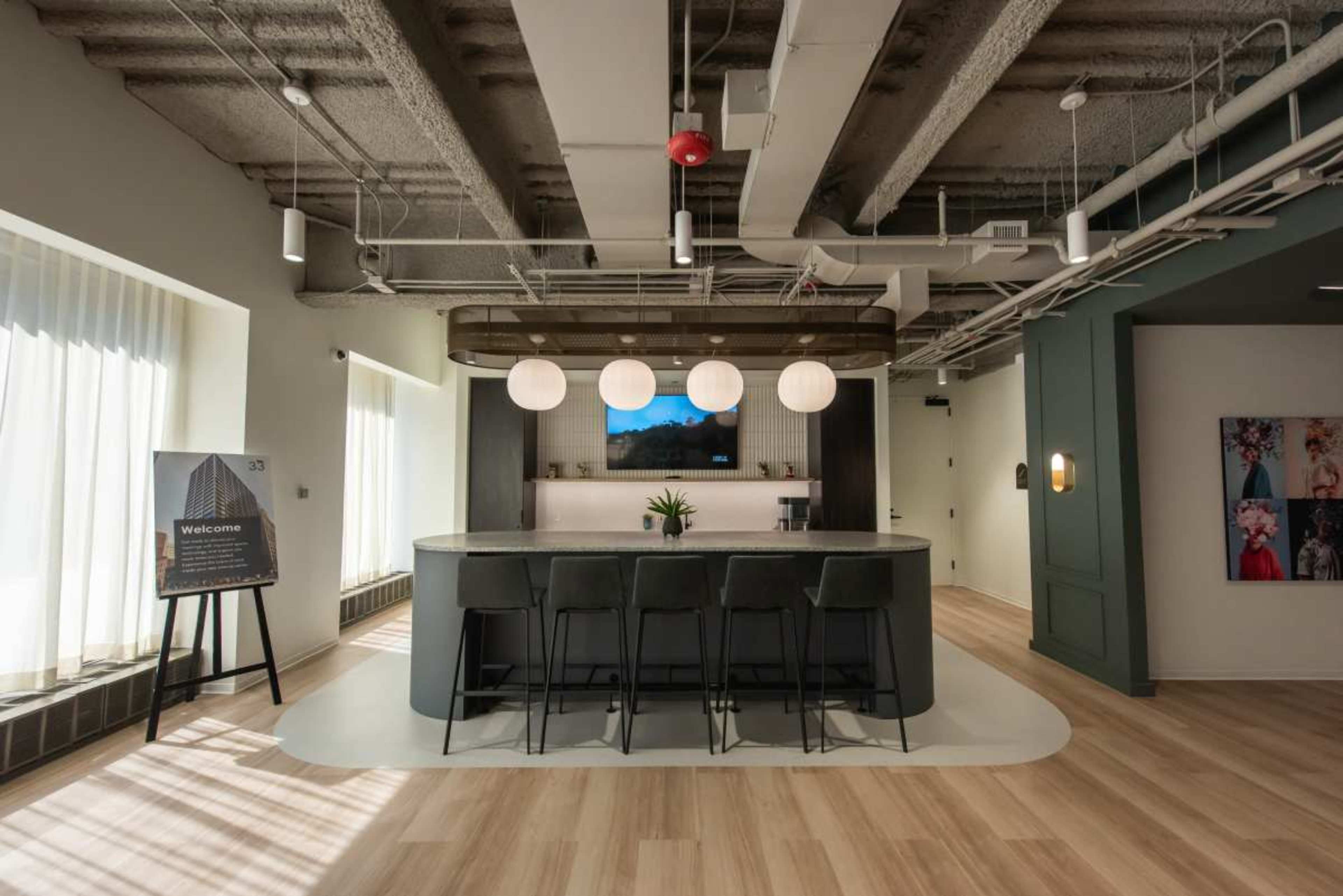 The image shows a modern office reception area featuring a curved bar with high chairs, pendant lights, and a welcoming sign nearby.