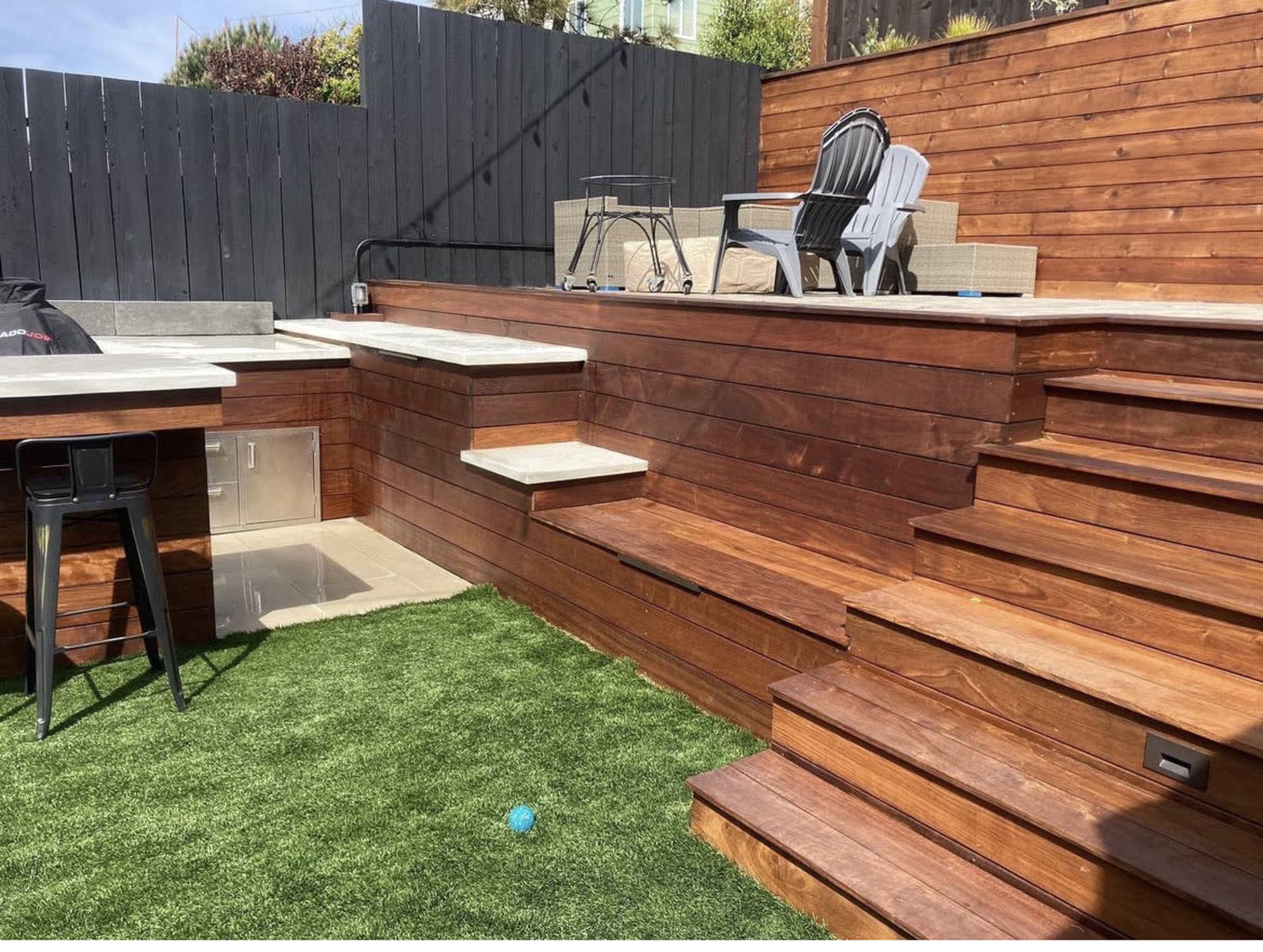 A backyard area featuring wooden steps leading up to a tiered deck with seating and landscaping made of artificial grass.