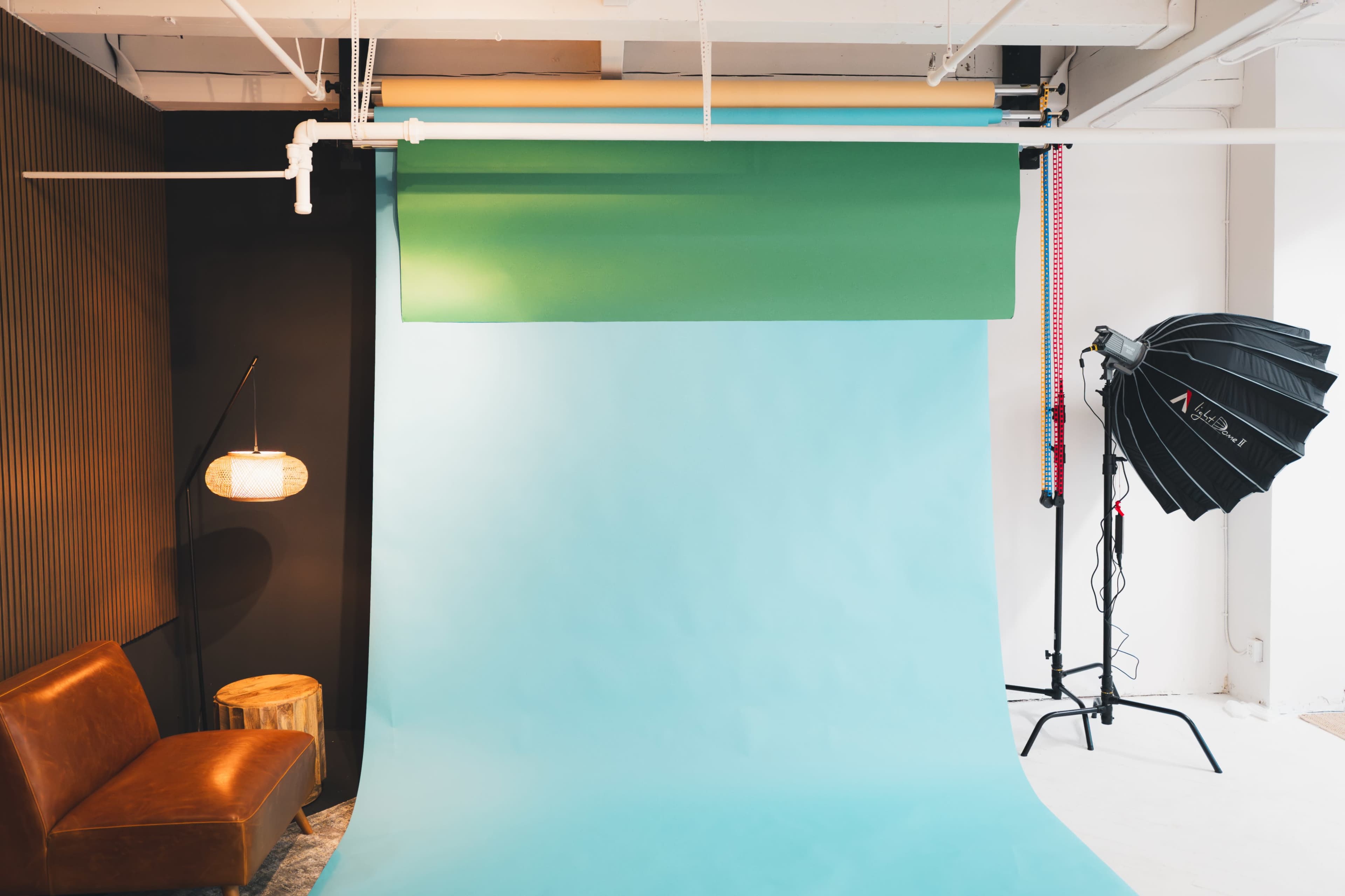 A photography studio setup featuring a light blue backdrop, a green roll-up paper backdrop, a softbox light, and a brown leather sofa against a wall.